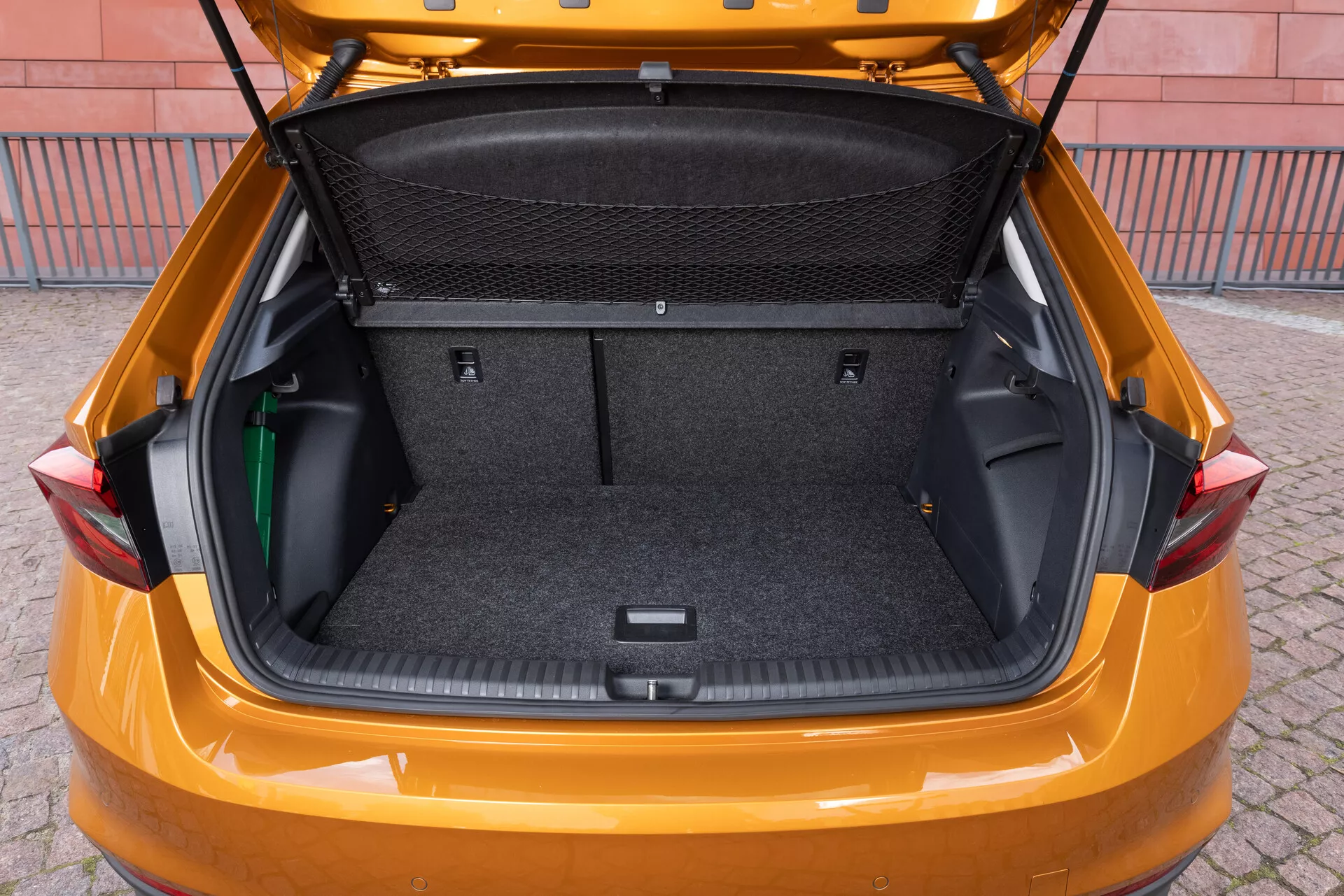 Slika prikazuje prtljažnik modela Skoda Fabia iz leta 2021, posneta iz rahlo povišanega zadnjega kota, da pokaže prostorno nakladalno območje. Notranjost je obdana s preprogastim materialom in vsebuje prilagodljive stranske shranjevalne prostore, tovorno mrežo in vdolbino na spodnjem delu za dodatno shranjevanje. Prtljažna vrata so dvignjena, kar razkriva velik čist prtljažnik, zasnovan tako, da poudarja skladiščne zmogljivosti in praktičnost vozila.