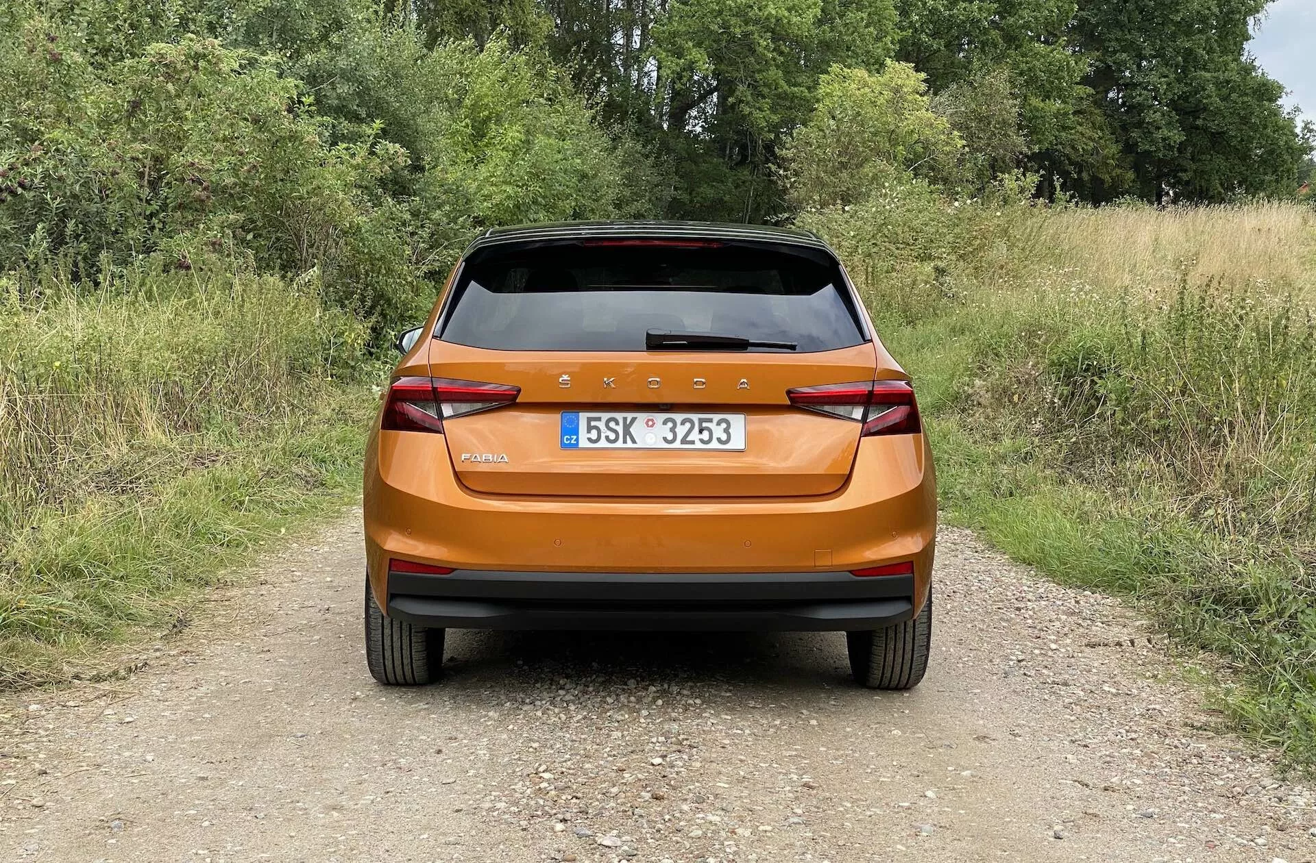 Zadnji pogled na svetlo kovinsko oranžnega hatchbacka Škoda Fabia 2021, fotografiranega podnevi na ozki zemljiščni poti v podeželskem okolju. Slika zajema sodoben hrbtni del z neprekinjeno zadnjo svetlobno vrstico, ostrimi kotnimi zadnjimi lučmi, vgrajenimi odsevniki, brisalnikom zadnjega stekla in zadnjim spojlerjem za aerodinamičnost, poudarja svetlečo površino in praktične lastnosti avtomobila.