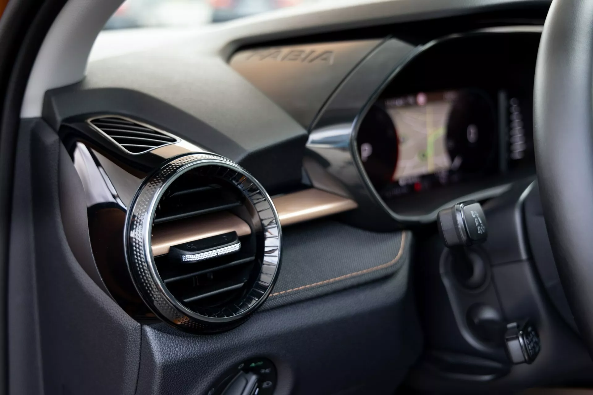 Podroben pogled notranjosti vozila Škoda Fabia 2021, osredotočen na stran sopotnika. Slika zajema okrogli zračni ventil z kovinsko ali kromirano površino na plošči s teksturo iz ogljikovih vlaken, kar kaže na športno ali prestižno opremo. Na desni je viden rob zaslona na dotik za infotainment, vgrajenega v armaturno ploščo. Pod zračnim ventilom so različni kontrolni gumbi, verjetno za avdio sistem, klimatizacijo ali druge funkcije vozila. Mala globinska ostrina poudari zračni ventil, obenem pa rahlo nakaže okoliške tehnološke komponente.