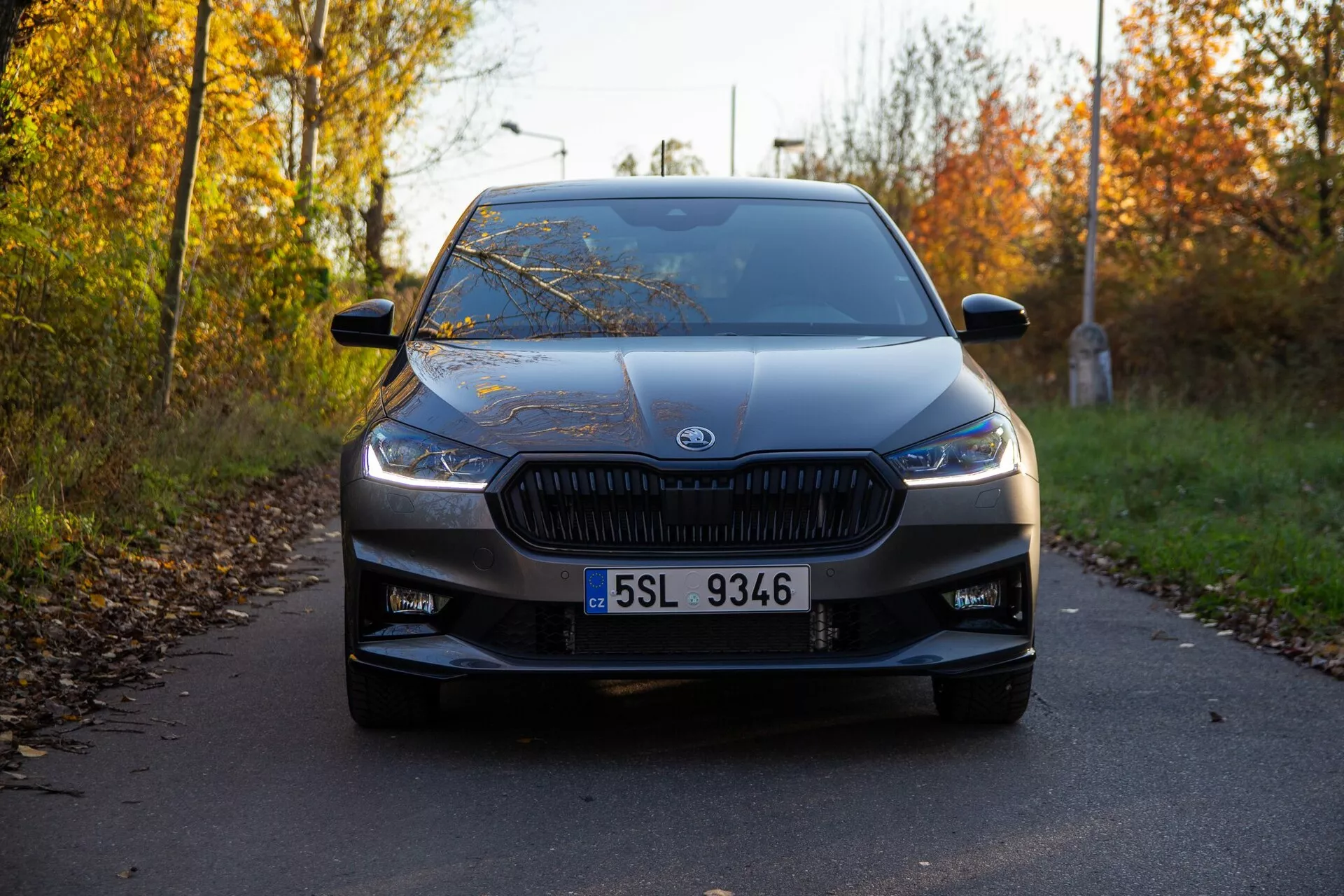 Sprednji pogled na Skoda Fabia 2021, posnet z nizkega kota, poudarja značilno rešetko z navpičnimi rebri in moderne, ozke žaromete. Avto, ki je centriran v okvirju, je parkiran na ozki cesti, ki jo obdaja jesensko listje, z jasno vidnim emblema proizvajalca, nameščenim na sredini rešetke. Vidni so tudi drugi oblikovalski elementi, kot je jasna registrska tablica, zračne odprtine na spodnjem odbijaču, nežne obrisne linije na pokrovu motorja, široko vetrobransko steklo in vzratna ogledala z indikatorji. Mehka svetloba in dolge sence nakazujejo, da je bila slika posneta bodisi zgodaj zjutraj ali pozno popoldne, pri čemer avto izgleda v odličnem stanju.