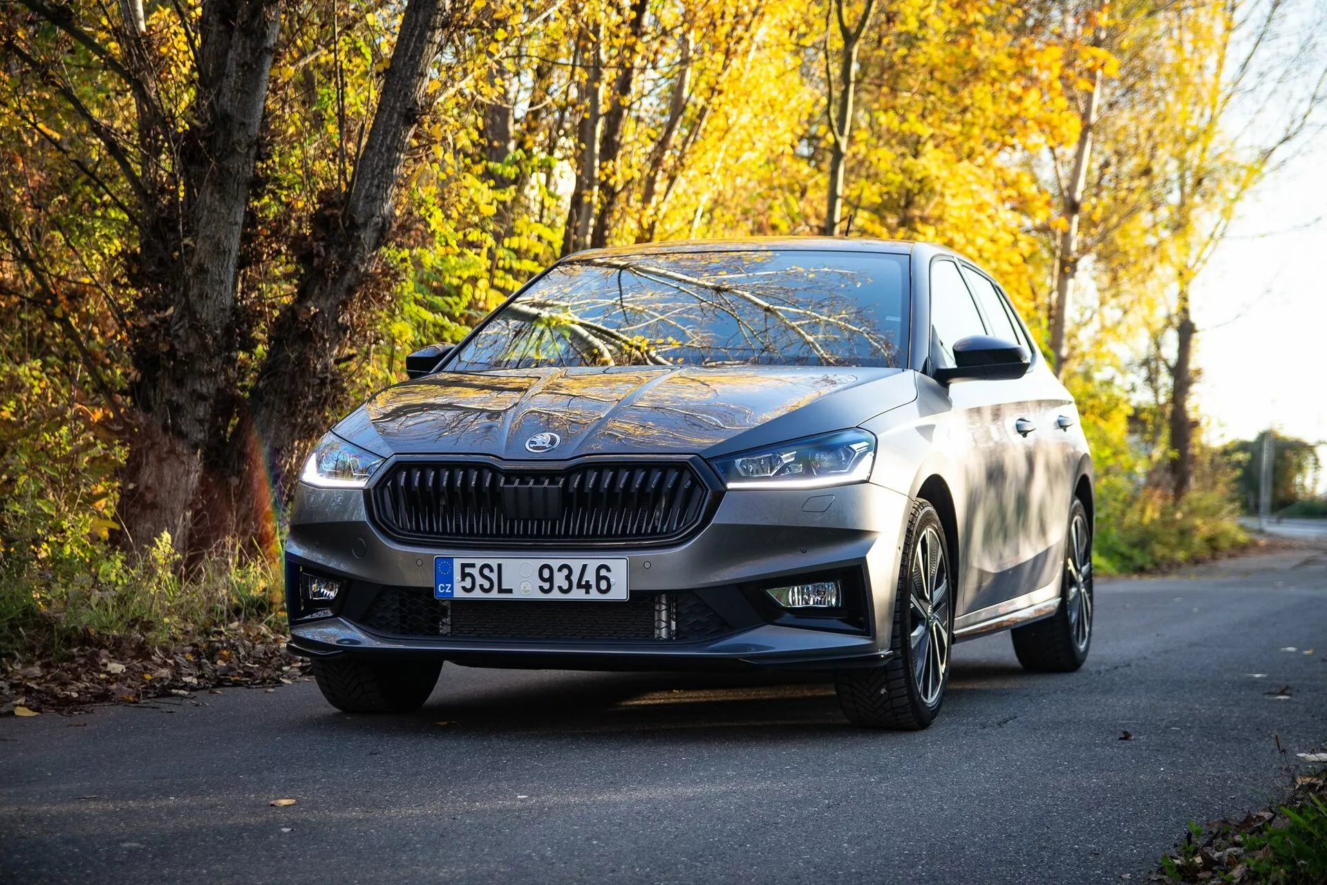 Slika modernega sedana Skoda Fabia 2021, parkiranega na cesti, obkroženi z jesenskim listjem, posneta iz perspektive na tleh, poudarja sprednjo mrežo, kotne žaromete, elegantne oblikovalske linije, litkolesarska platišča in stranska ogledala z vgrajenimi smerniki, ki odražajo visoko kakovost kovinske barve in kombinacijo luksuza in športnosti.