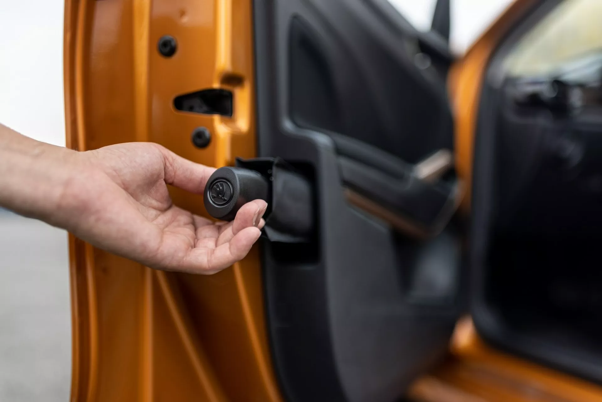 Od blizu fotografija voznikovih vrat vozila Skoda Fabia 2021, rahlo priprta, gledana od zunaj z vidnim notranjim robom. Roka drži kompakten, črn, okrogel ključek, ki nakazuje brezključni vstop ali avtomobilski varnostni sistem. Avto je svetlo oranžne barve z kovinskim leskom, prikazuje napredne mehanizme zaklepanja in prilagojene varnostne funkcije.
