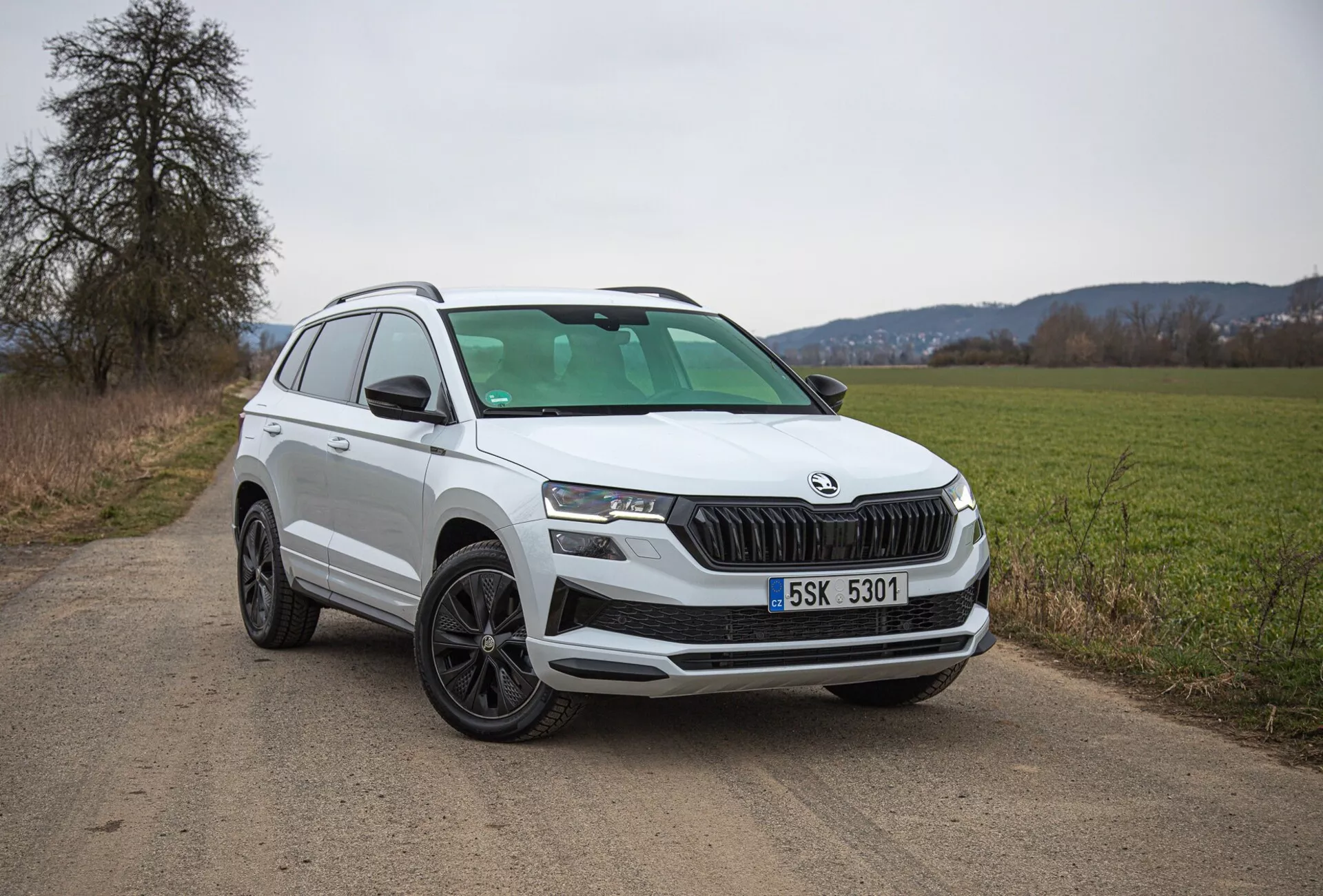 Beli SUV Škoda Karoq 2021 parkiran na podeželski makadamski cesti, prikazuje pogled na četrtino spredaj levo, vključno z rešetko, žarometi in oblikovanjem koles, s poudarkom na sodobnih značilnostih, kot sta LED osvetlitev in parkirni senzorji.