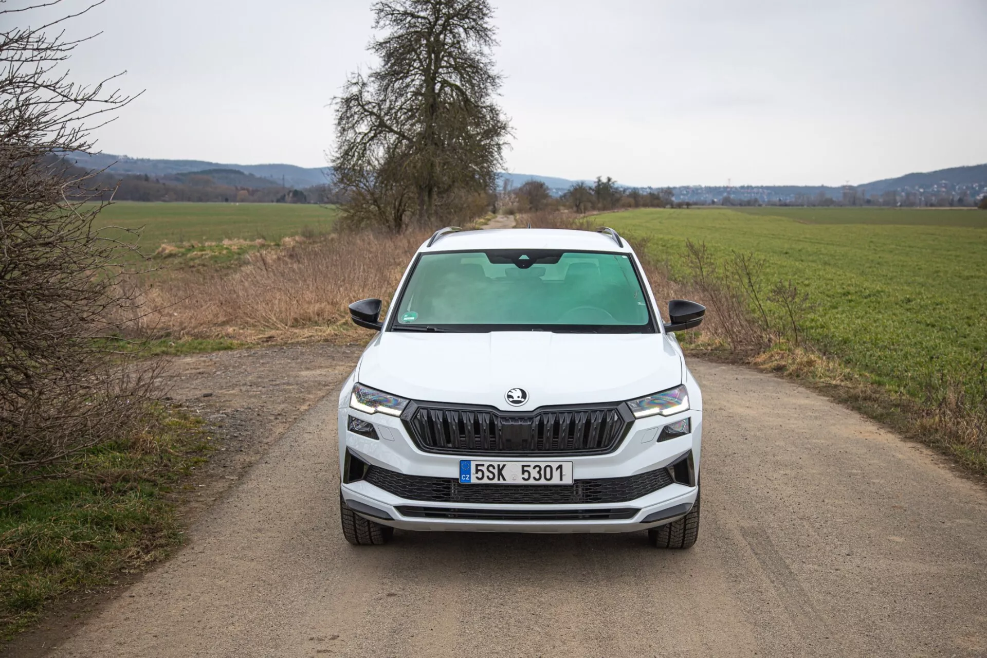 SUV Škoda Karoq 2021 prikazuje neposreden sprednji pogled na ozko, neasfaltirano podeželsko cesto, poudarja značilno rešetko Škoda, ostre LED žaromete, meglenke znotraj črnega okvira za potencialne terenske sposobnosti, dinamične linije pokrova motorja in napredne tehnološke značilnosti na prednjem delu vozila.