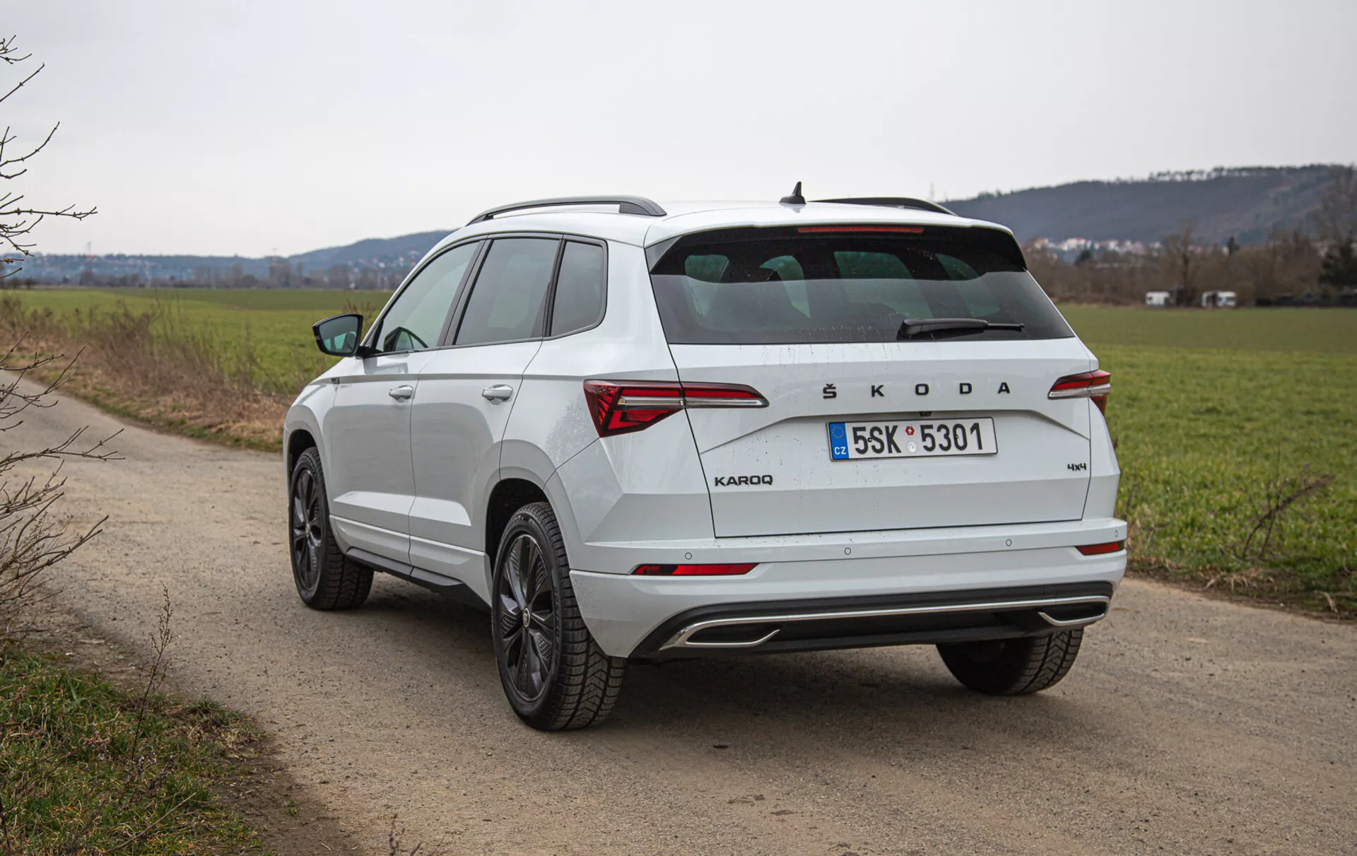 Zadnji pogled iz treh četrtin na beli kompaktni SUV Škoda Karoq na podeželski cesti, s poševnim zadnjim oknom, strešnimi nosilci, LED zadnjimi lučmi, blagovno znamko Škoda, večkrakimi litimi platišči in kromiranimi detajli, fotografirano na prostem podnevi z naravnim krajinskim ozadjem.