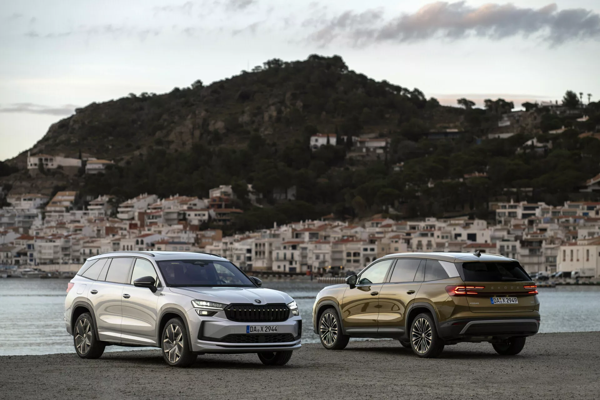 SUV Skoda Kodiaq 2024 parkirana drug ob drugem v zori na obalni promenadi z ozadjem obalnega mesta, prikazuje sprednjo mrežo, žaromete in elegantne oblikovne linije enega in zadnji tričetrtinski pogled drugega.
