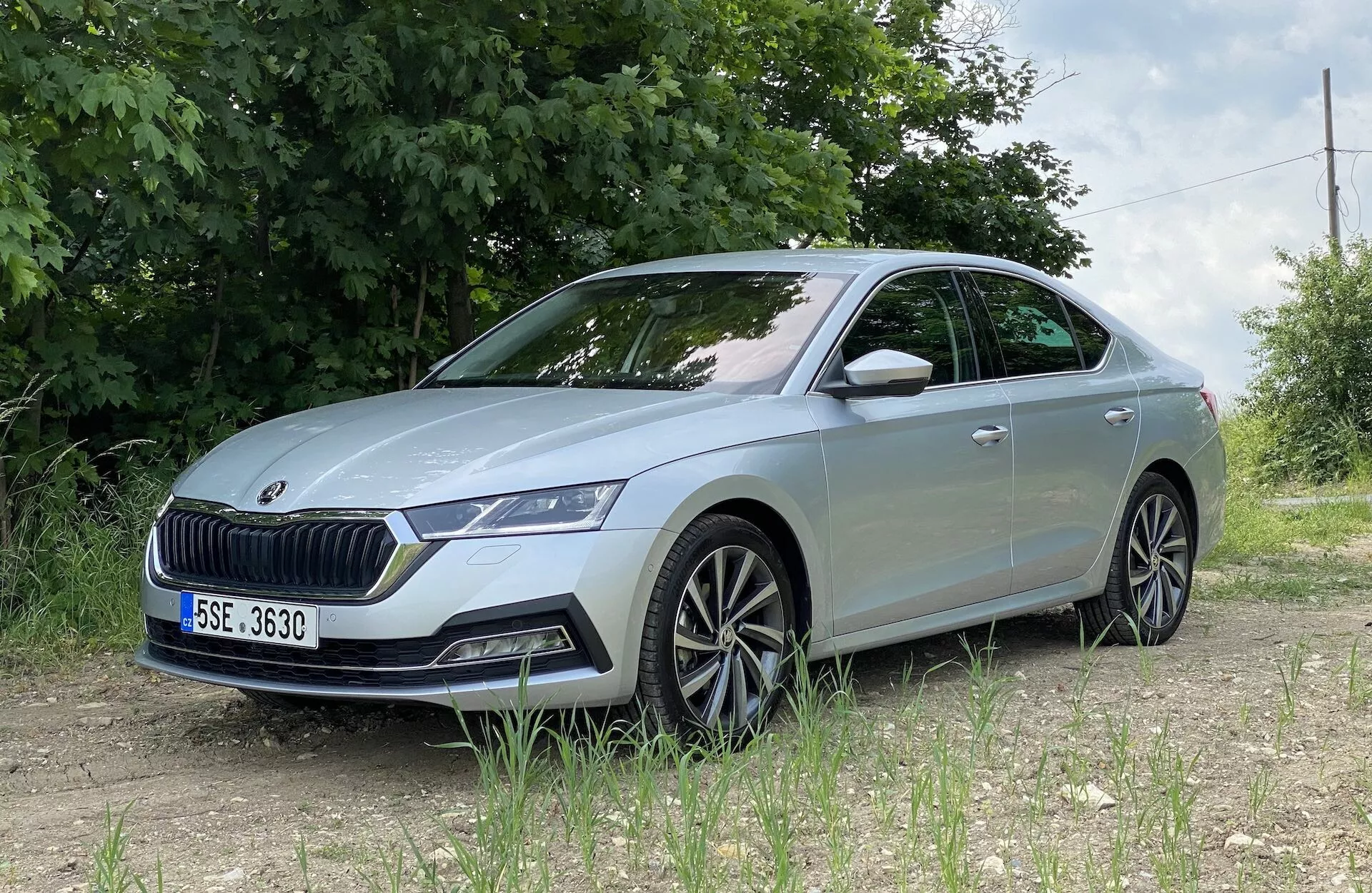 Skoda Octavia 2020, gledana iz nizkega sprednjega tričetrtinskega kota, prikazujoc podrobni sprednji levi del, vključno z enoto LED žarometov, prominentno mrežo, sprednjim odbijačem z senzorji, kiparskimi linijami pokrova motorja, večnaperjalnih platišč, vgrajenim smernikom na stranskem ogledalu in rahlo zatemnjenimi okni, pozicionirana na neasfaltirani površini z zelenim listjem in žično ograjo v podeželskem ali pol-podeželskem okolju.