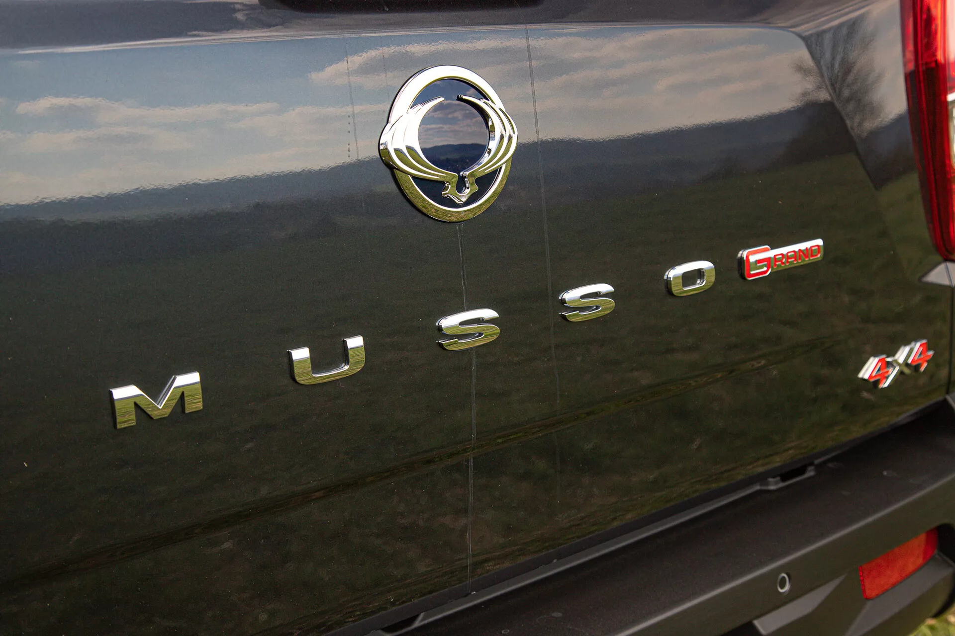 Podroben pogled na zadnji del SsangYong Musso Grand iz leta 2022, ki prikazuje kromirano napis ‘MUSSO’, srebrni emblem proizvajalca z stiliziranim motivom ptica in rdečo oznako ‘E-XDI Sport’, ki označuje tip motorja in raven zmogljivosti, postavljeno proti temni, verjetno črni, zunanji strani vozila z odsevnimi kromiranimi in teksturiranimi podrobnostmi.