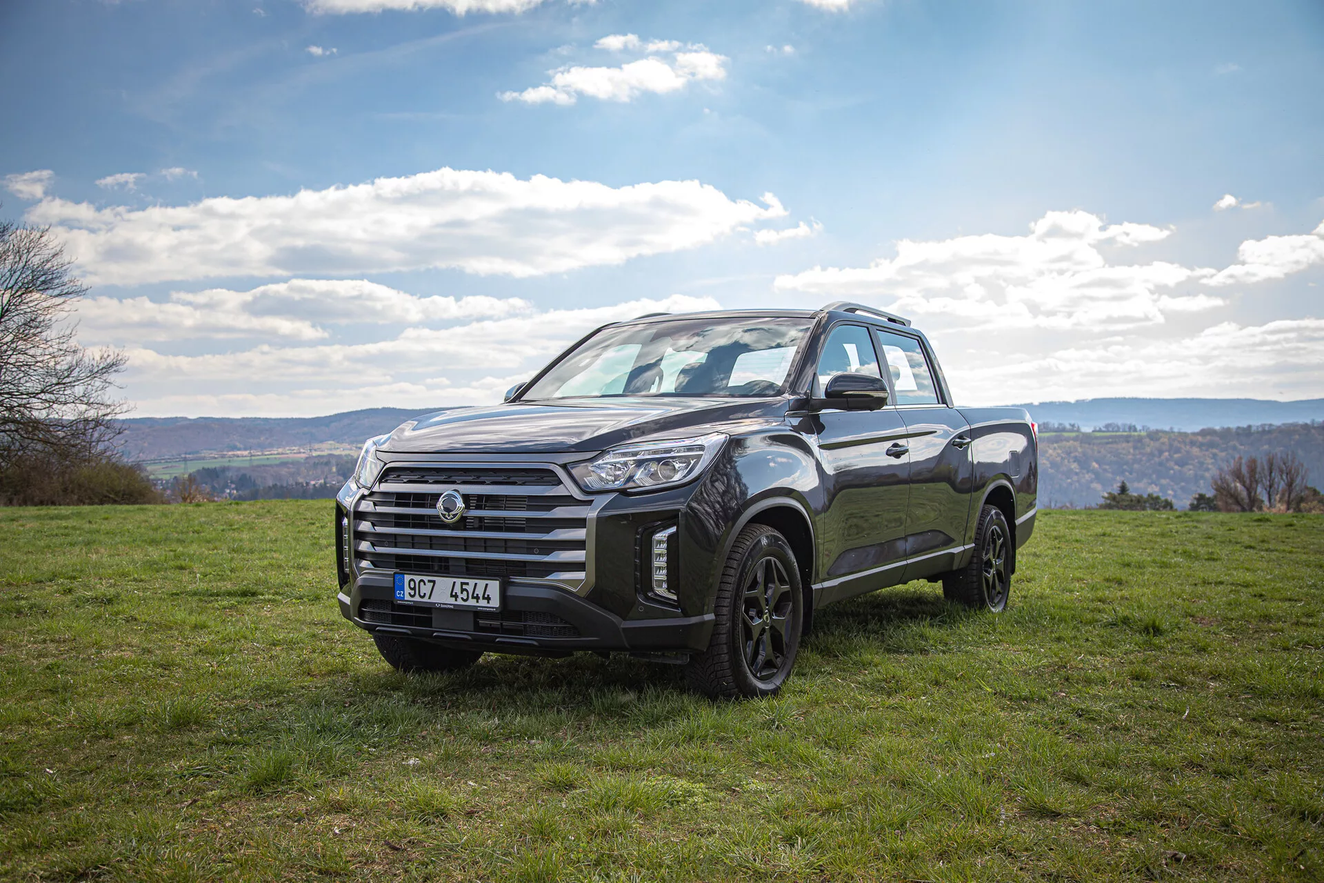 Pick-up SsangYong Musso Grand iz leta 2022 parkiran na travnati pokrajini pod oblačnim nebom, prikazuje svojo robustno zasnovo in sposobnost terenske vožnje s sprednjega pogleda tri četrtine med dnevno svetlobo.