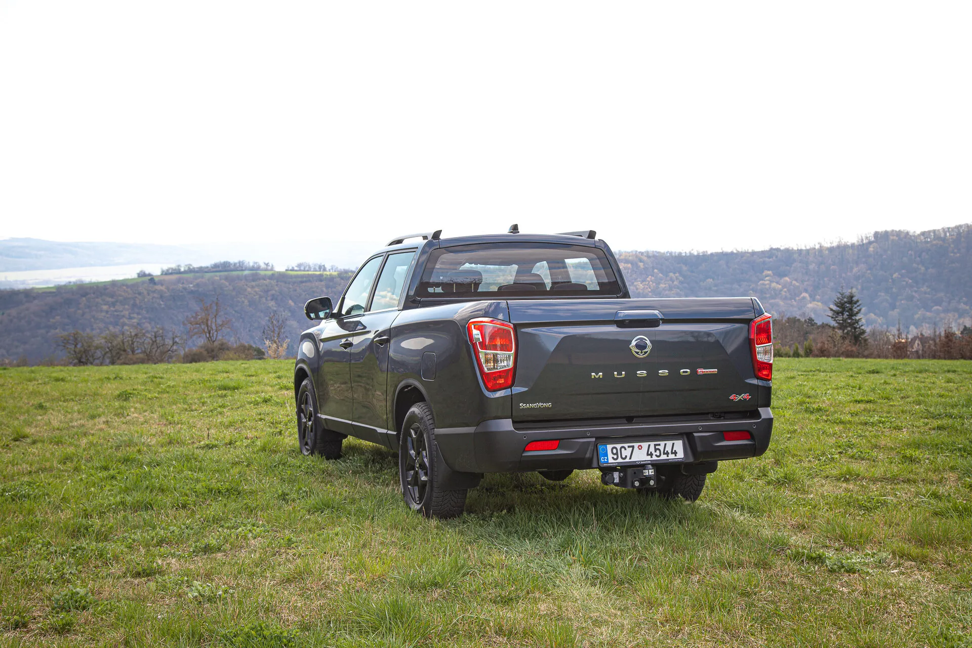 Pickup SsangYong Musso Grand letnik 2022, posnet z zadnjega kota, ki poudarja športno-utilitarni videz, štirivratni dizajn in vsestranskost na prostem ob slikovitem hribovitem in morda rečnem ozadju podnevi
