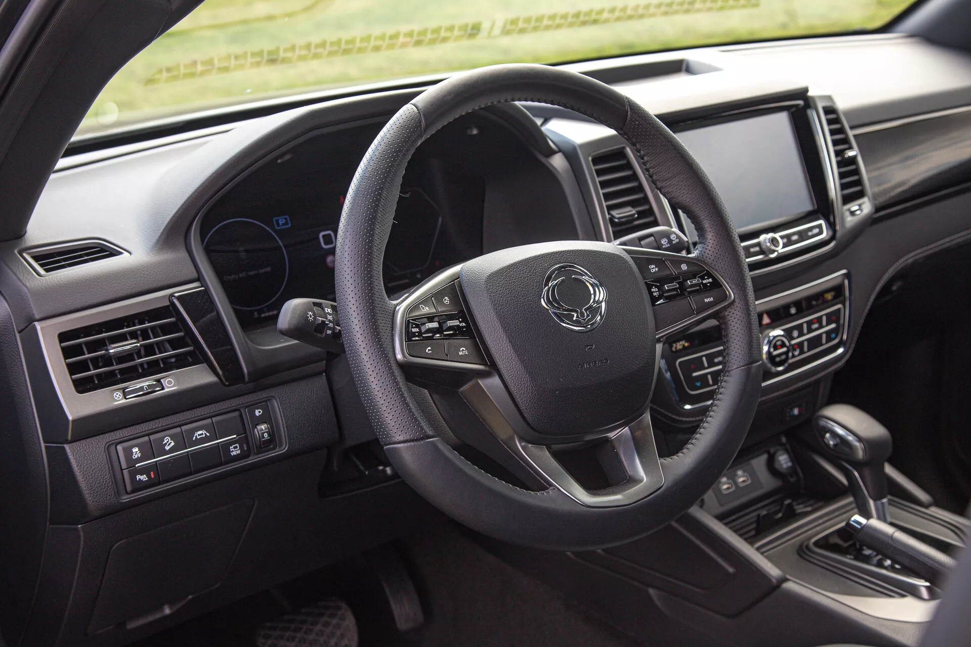 Posnetek notranjosti SsangYong Musso 2022, ki prikazuje stran voznika, vključno s sodobnim večfunkcijskim volanom s tremi kraki, digitalno instrumentno ploščo in centralnim infotainment sistemom na armaturni plošči iz perspektive zadnjega potnika