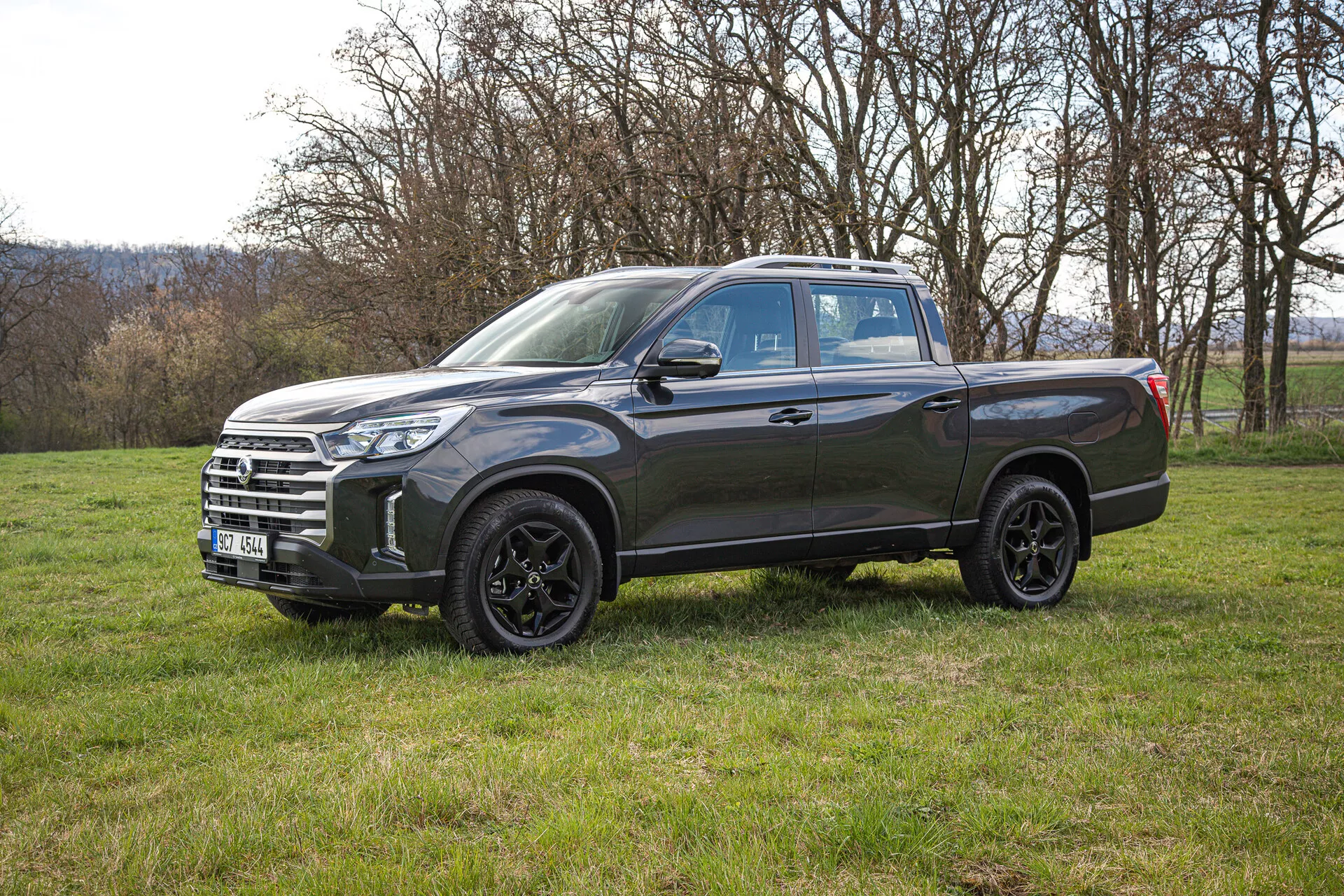 Stranski pogled na temno siv pickup SsangYong Musso Grand 2022 na travniku, prikazuje njegova lita kolesa, LED žaromete in prostorno tovorno površino