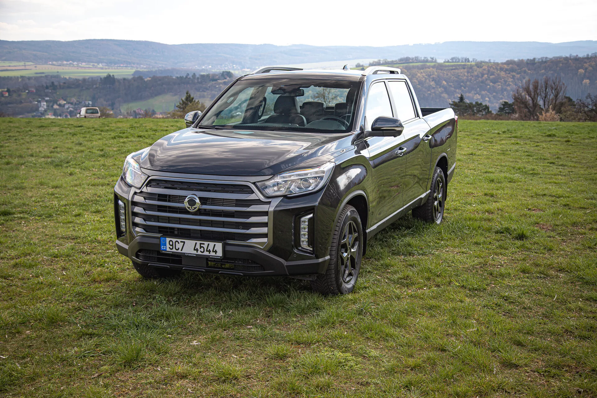 Pickup SsangYong Musso Grand 2022 v mirujočem položaju na travnatem pobočju prikazuje sprednji in bočni pogled vključno z veliko masko, kotnimi svetlobnimi sklopi in zasnovo koles primernih za vožnjo izven cest, z ruralno krajino v ozadju.
