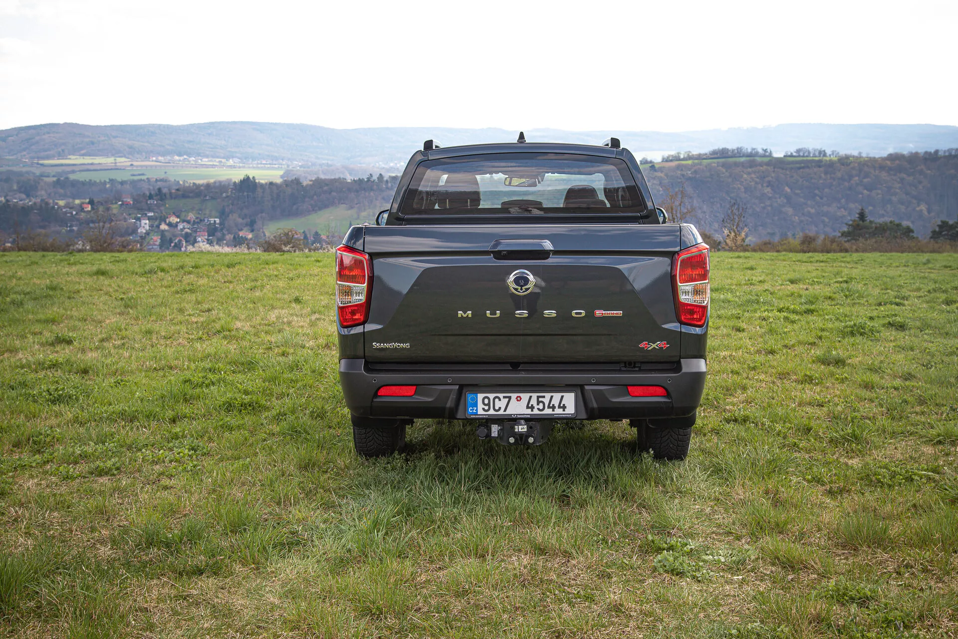 Zadnji pogled na temni pickup SsangYong Musso Grand 2022 na neenakomernem travnatem terenu z ruralno pokrajino v ozadju, prikazuje branding na prtljažnih vratih, vertikalne zadnje luči, zadnje senzorje in vlečno kljuko podnevi