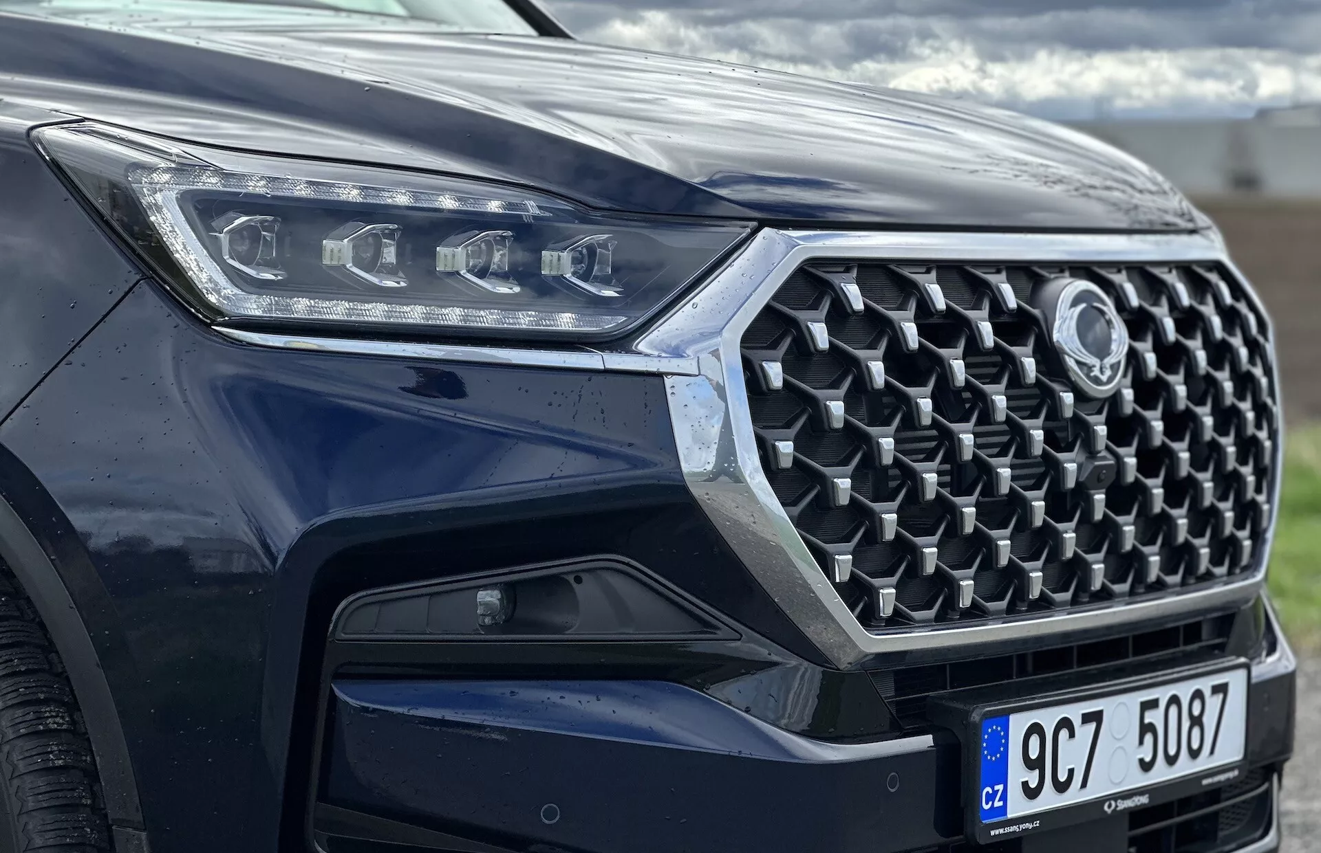 Pogled spredaj pod kotom na SsangYong Rexton 2021, osredotočen na rešetko, sprednji odbijač in dizajn LED žarometov, ki prikazuje napredne varnostne funkcije in sodobno tehnologijo z metalik barvo.
