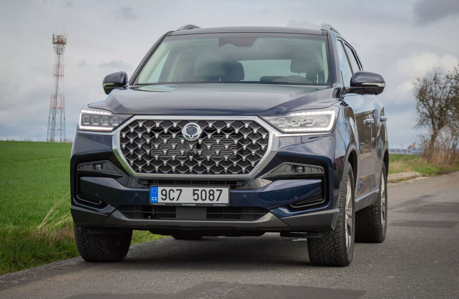 SUV SsangYong Rexton 2021 na podeželski cesti, prikazuje podrobni pogled na njegov sprednji dizajn z geometrijsko rešetko in LED žarometi, ki odraža napredno tehnologijo in terenske sposobnosti
