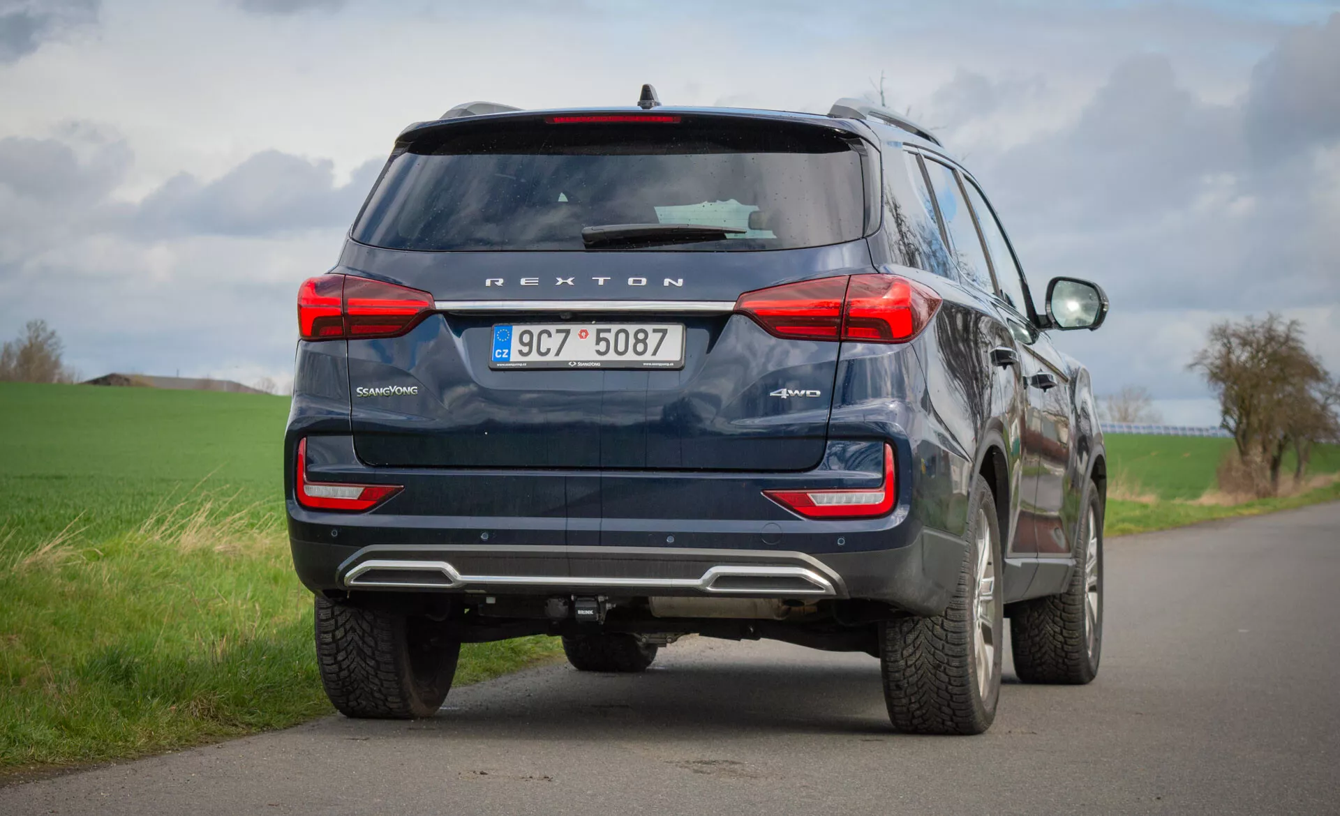 Zadnji pogled na modri SUV SsangYong Rexton 2021, parkiran na podeželski cesti z dvema pasovoma, ki poudarja njegove oblikovne značilnosti, kot so zadnje luči, zadnje okno, izpušne cevi in varnostna tehnologija, vključno s kamero za vzvratno vožnjo in parkirnimi senzorji pod oblačnim nebom.