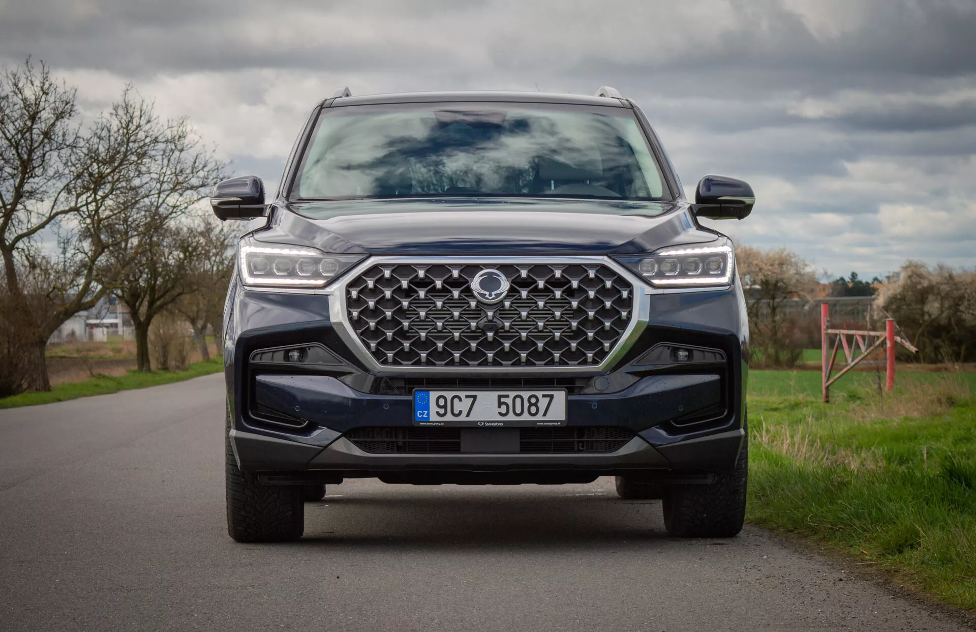 Moderen SUV SsangYong Rexton 2021 v barvi, posnet iz klečečega položaja, ki prikazuje sprednji del vozila, LED žaromete, veliko oblikovano rešetko, sprednji odbijač s podložko za drsenje in napredne sisteme pomoči vozniku