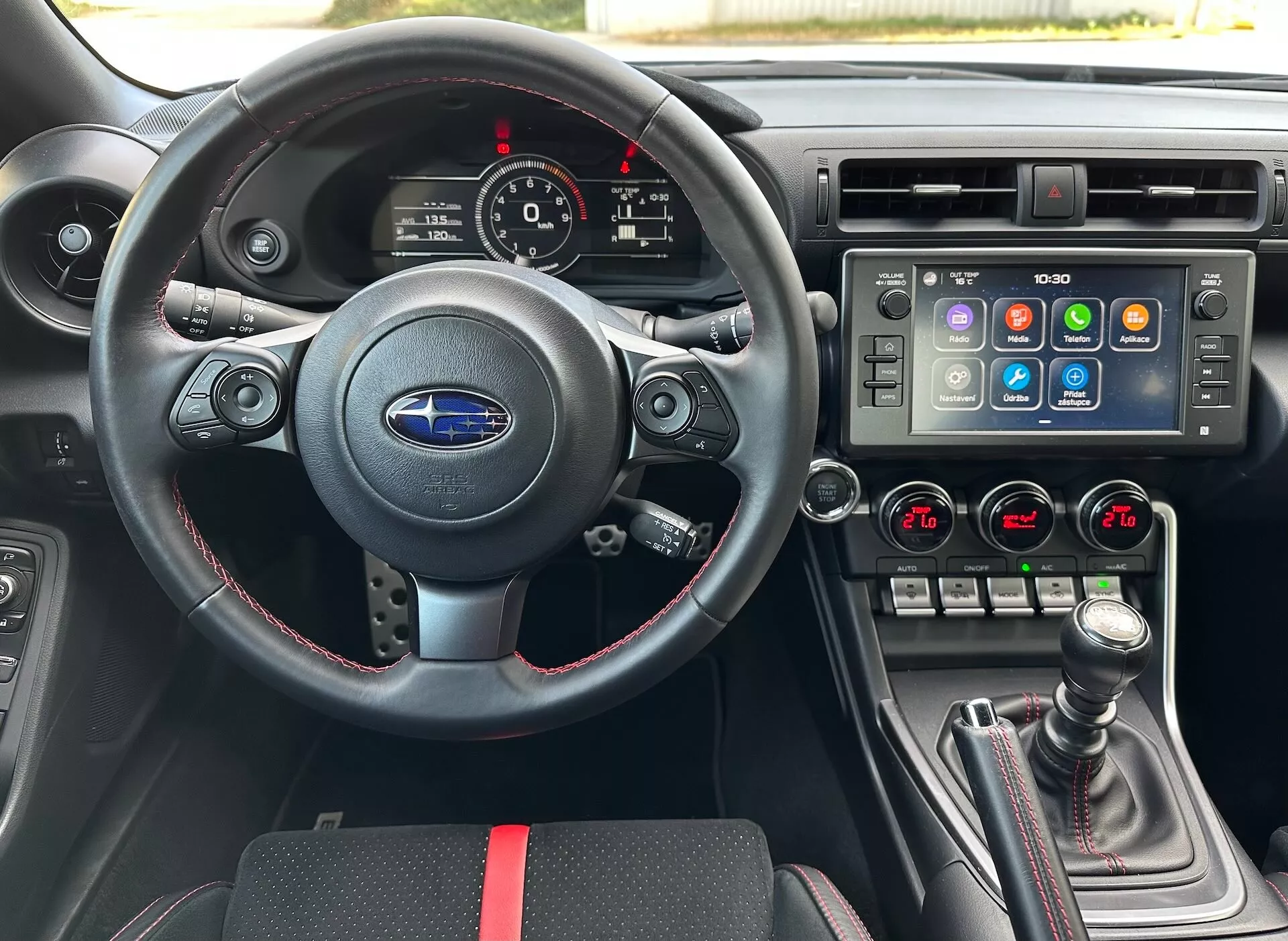 Notranjost Subaru BRZ 2023 iz perspektive voznika, ki vključuje športni usnjen volan z večfunkcijskimi kontrolami in rdečimi šivi, instrumentno ploščo z analognimi številčnicami in digitalnim zaslonom, osrednjo konzolo z velikim zaslonom na dotik za informacijsko zabavni sistem in dvopodročno klimatsko kontrolo, in ročni menjalnik z usnjenim zaščitnim pokrovom in rdečimi poudarki, proti črni notranjosti s športnimi rdečimi poudarki, kar poudarja napredne funkcije vozila in tehnologijo.