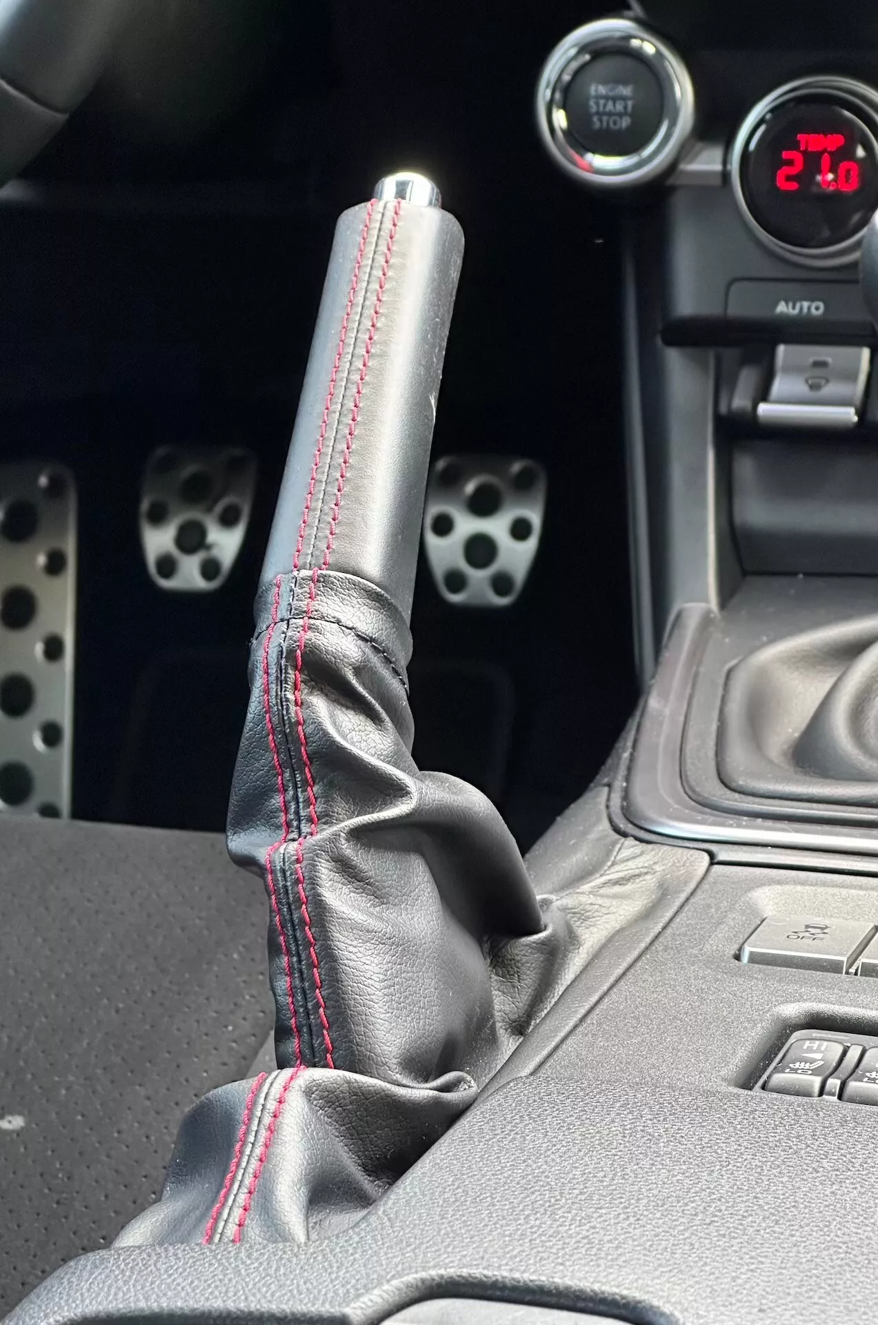 Podrobni posnetek sredinske konzole Subaru BRZ 2023, ki poudarja kovinsko prestavno ročico z rdečim kontrastnim šivom na usnjenem čevlju, digitalni prikazovalnik, ki prikazuje '21', športne aluminijaste pedale z vrtanimi luknjami in tehnološke funkcije kot so gumbi za elektronski stabilnostni nadzor.