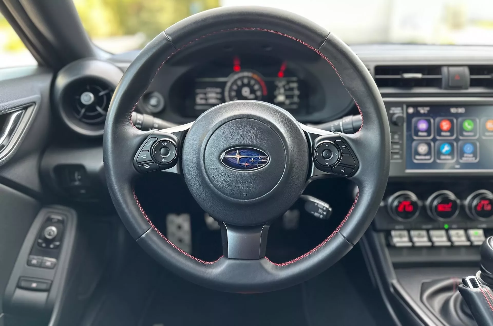 Notranjost Subaru BRZ 2023 iz perspektive voznika, osredotočena na triskladi volan z rdečim šivanjem, logotip Subaru, armaturo z merilniki in sredinsko konzolo