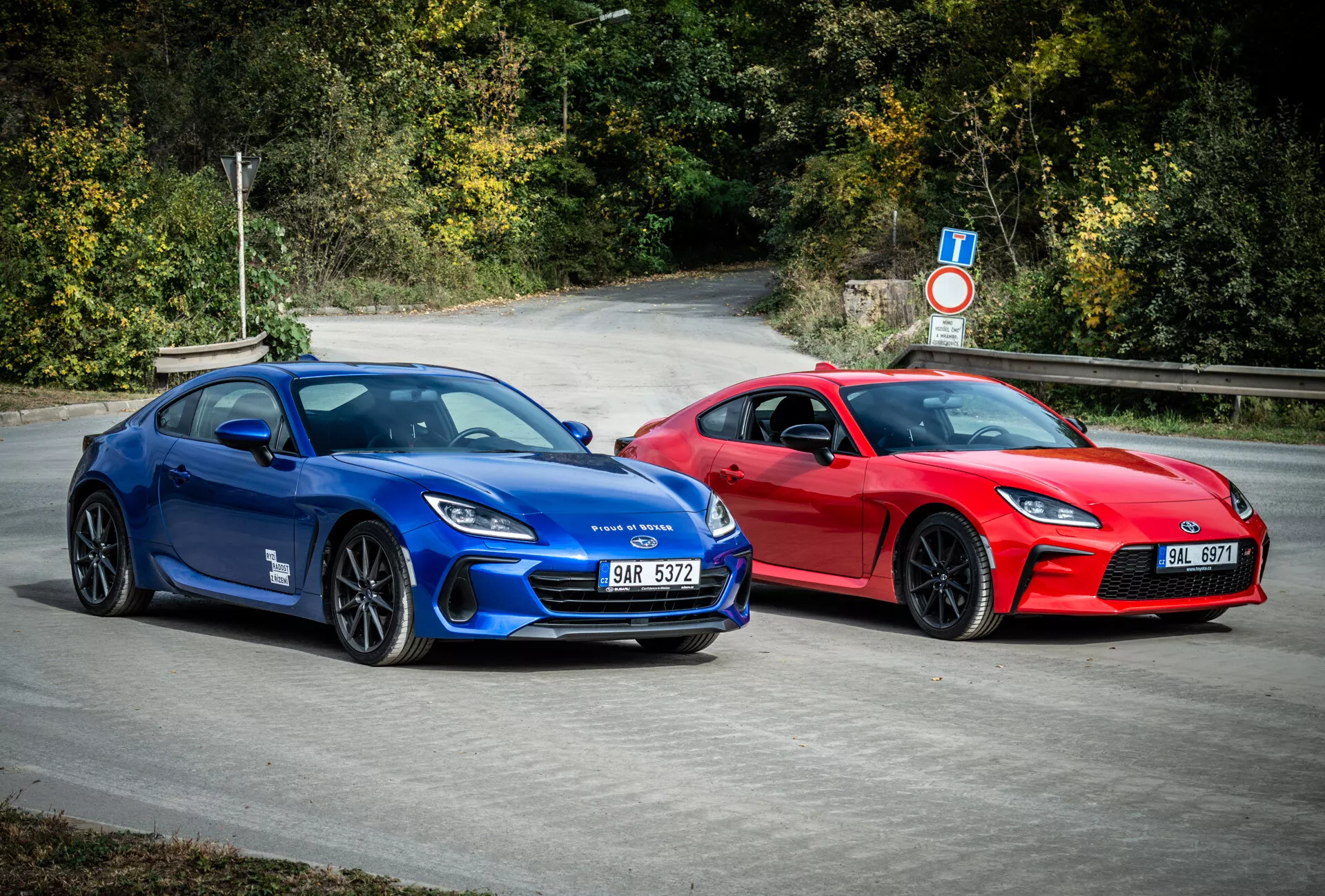 Športni kupéji Subaru BRZ 2023 v modri in rdeči barvi z aerodinamičnim dizajnom in zmogljivostnimi lastnostmi, parkirani na prostem, obdani z naravno listnato okolico