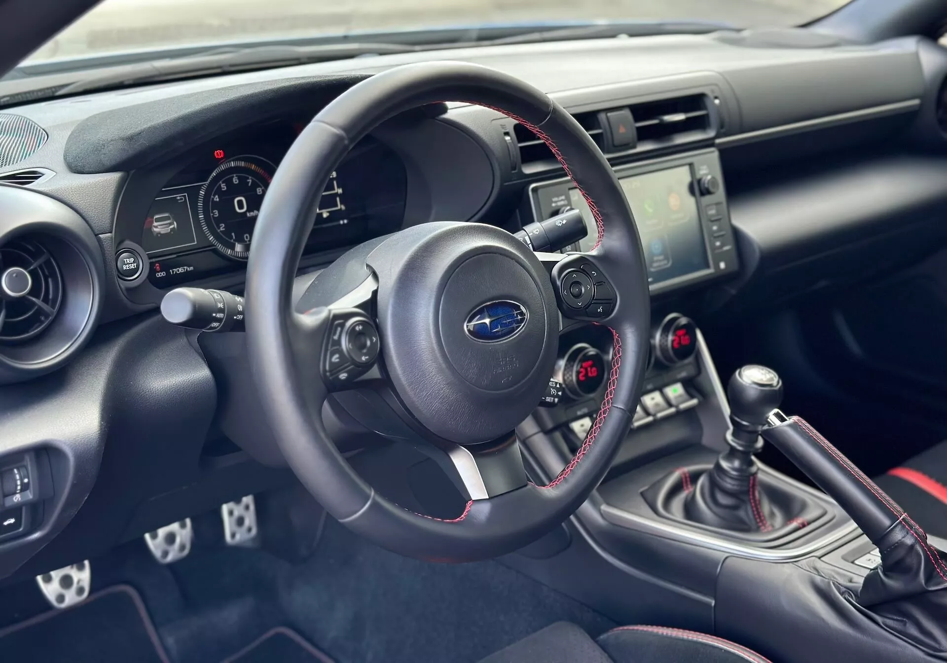 Notranjost Subaru BRZ 2023 osredotočena na armaturno ploščo in kokpit na strani voznika z detajlnim prikazom volana, instrumentne plošče, sredinske konzole z nadzorom za avdio in klimo, ter ročno prestavno ročico s športnimi rdečimi šivi