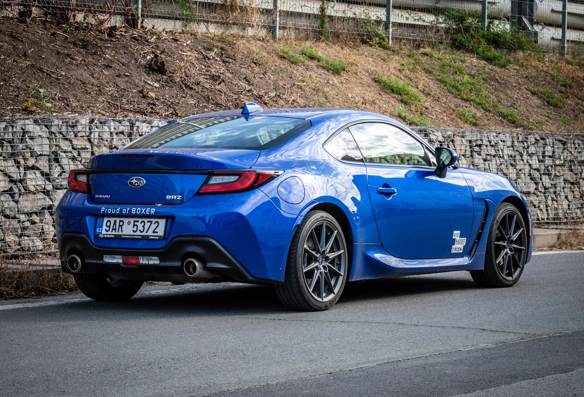 Športni kupe Subaru BRZ letnik 2023 v metalik modri barvi, parkiran na naklonjeni cesti, ki prikazuje zadnji tričetrtinski pogled vozila, poudarjajoč bočni profil vozila, ukrivljenost zadnjega dela, športna lita platišča, dvojne izpušne konice, spojler na pokrovu prtljažnika in značko motorja 'Proud of Boxer'.