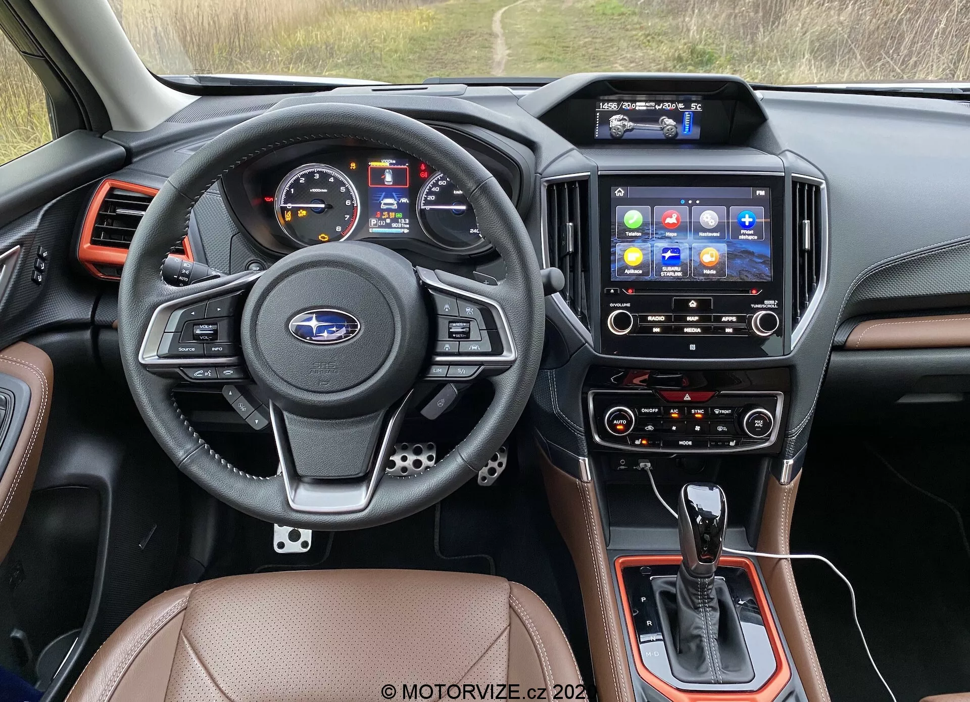 Posnetek notranjosti iz voznikovega sedeža modela Subaru Forester 2020, osredotočen na armaturno ploščo, volan, infotainment sistem in sredinsko konzolo. Usnjen volan s tremi kraki ima večfunkcijske kontrole za avdio, telefon, glasovne ukaze in tempomat, z emblemom Subaru na sredini. Instrumentna plošča vključuje digitalni zaslon in analogne merilnike hitrosti in vrtljajev motorja. Sredinska konzola vsebuje prestavno ročico avtomatskega menjalnika, dodatne kontrole in morebiti izbirnik načina vožnje. Infotainment zaslon na dotik z različnimi ikonami aplikacij ponuja navigacijo, medije, telefonsko povezljivost in nastavitve vozila. Spodaj so klimatske kontrole, ki verjetno omogočajo dvopodročno nastavitev za voznika in sovoznika. Notranjost je obdelana z usnjenimi ali umetnimi usnjenimi sedeži, črnimi plastičnimi komponentami in kovinskimi zaključki. Zračniki so postavljeni okoli infotainment sistema in na robovih armaturne plošče. Ta slika spada v kategorije notranjosti avtomobilov, avtomobilske tehnologije, armaturne plošče, infotainment sistemi in klimatske kontrole.