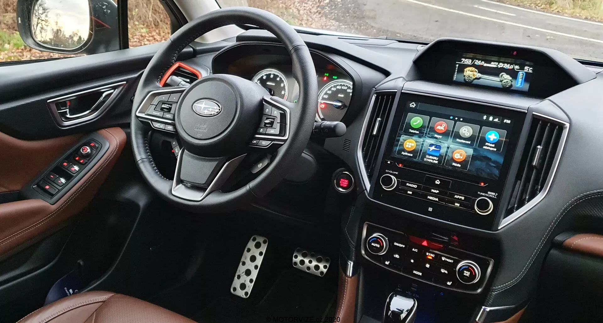 Pogled v notranjost Subaru Forester 2020 s sedeža voznika, prikazuje volan z nameščenimi krmilami, delno viden instrumentni grozd z analognimi merilniki in osrednjim digitalnim zaslonom, izrazit zaslon na dotik z barvnimi ikonami, ročne gumbe za nadzor klime, sedeže iz usnja in kovinske pokrove pedal.