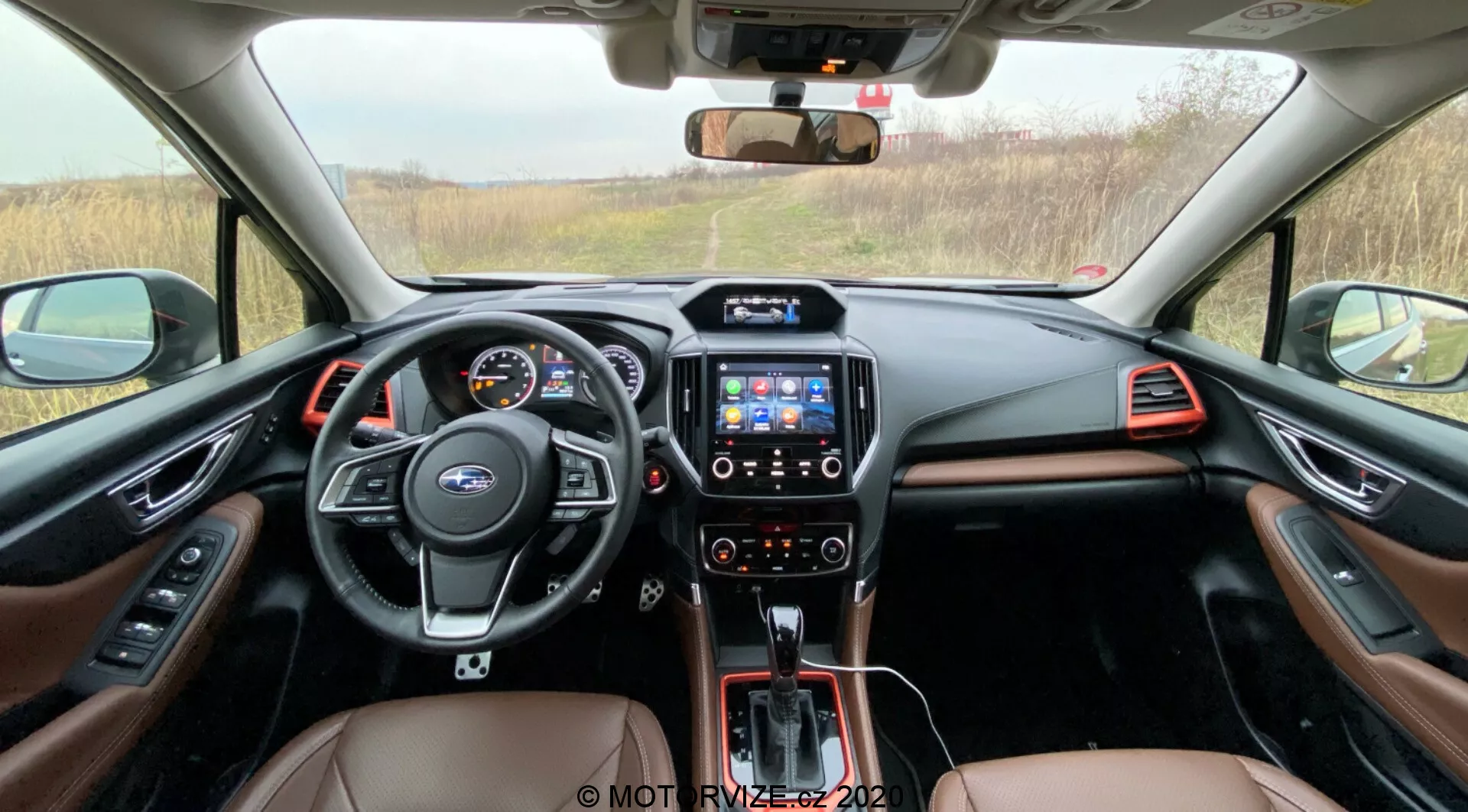 Notranji pogled na kabino Subaru Forester 2020, ki prikazuje večfunkcijski volan z upravljanjem, digitalno armaturno ploščo, sredinsko konzolo z infozabavnim sistemom na dotik, avtomatski menjalnik in dvobarvno notranjost z rjavim usnjem in kovinskimi okrasnimi elementi, vidno s sedežnega prostora nazaj proti armaturni plošči.