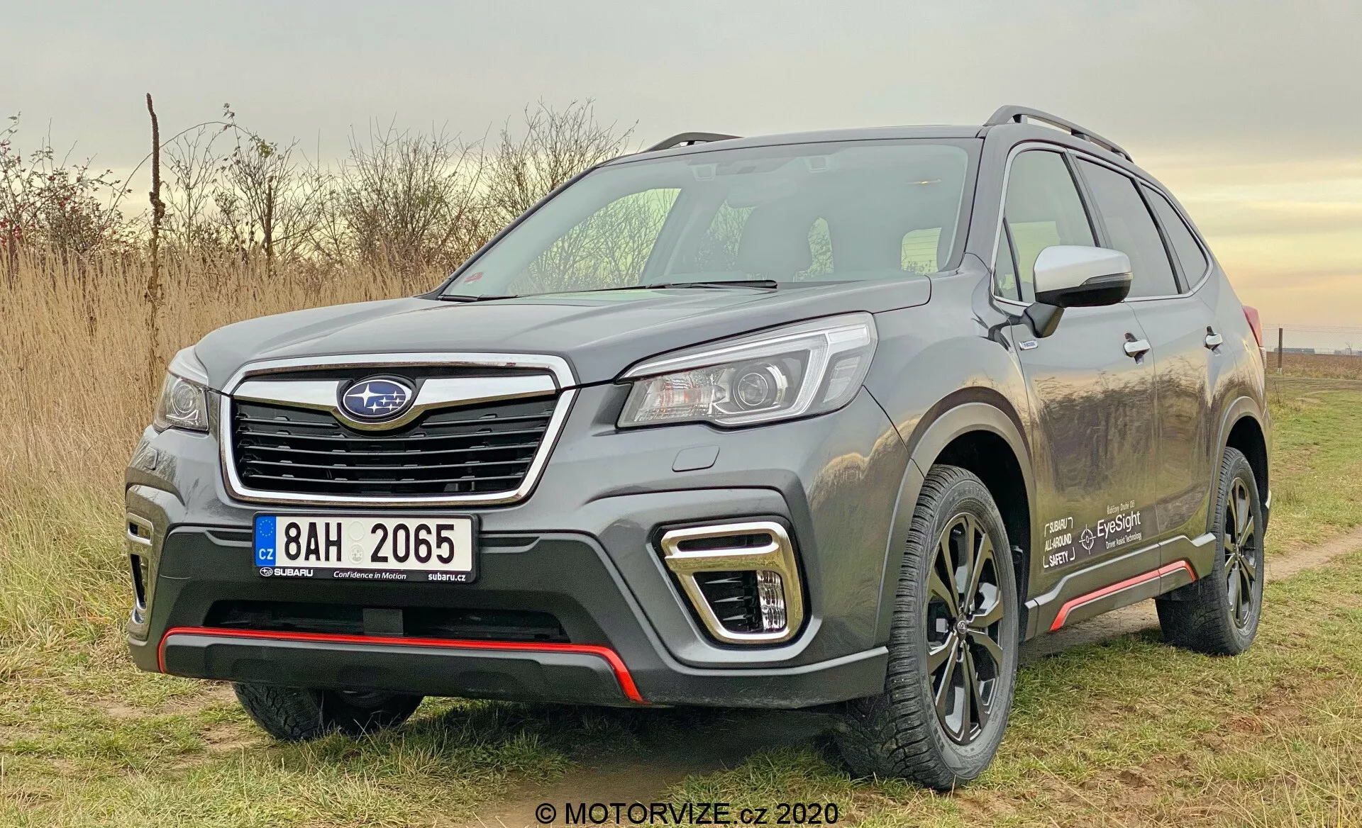 Slika prikazuje srebrno SUV Subaru Forester 2020, parkirano zunaj med dnevom v naravni svetlobi. Posneta je s sprednjega tričetrtinskega kota, fotografija jasno prikazuje levi sprednji del vozila, vključno s šesterokotno mrežo z emblemi proizvajalca, elegantne LED ali HID žaromete, dnevne luči v obliki črke C okoli meglenk in atletskega sprednjega odbijača s srebrnimi zaščitnimi elementi. Skulpturirana pokrov motorja ima dvignjene robove, nekoliko poudarjeni blatniki pa mu dajejo mišičast videz. Stranski profil vsebuje izrazito karakterno linijo, črne plastične obloge na spodnjem delu vrat, umazane aluminijaste platišča, strešne nosilce in delno vidno stransko ogledalo z vgrajenimi smerniki. Na sprednjem blatniku je značko, ki kaže na posebno raven opreme ali vrsto motorja. Okolje nakazuje lahkotno vožnjo po terenu, poudarjeno z brizganjem blata na straneh vozila.