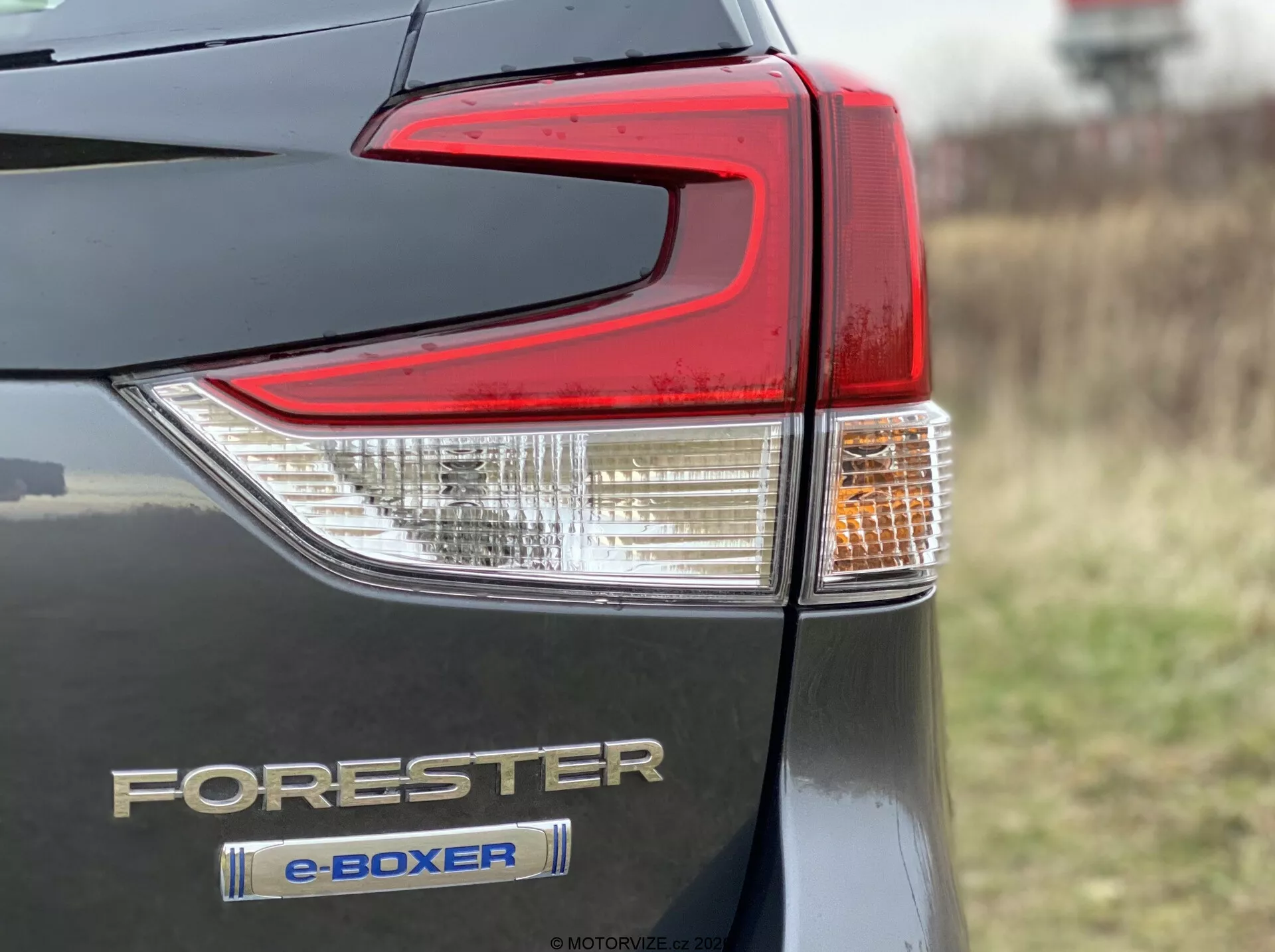 Podroben bližnji pogled na del zadnje luči na Subaru Foresterju 2020 z e-Boxer hibridno tehnologijo. Fotografija prikazuje zadnji levi kot z voznikovega pogleda, poudarja kompleksno oblikovanje zadnje luči s prozornimi in rdečimi lečnimi elementi, značke Forester in e-Boxer ter kovinsko karoserijo. Ostrina je osredotočena na značke in zadnjo luč, z zamegljenim ozadjem zaradi plitve globinske ostrine, posneto pod poševnim kotom.