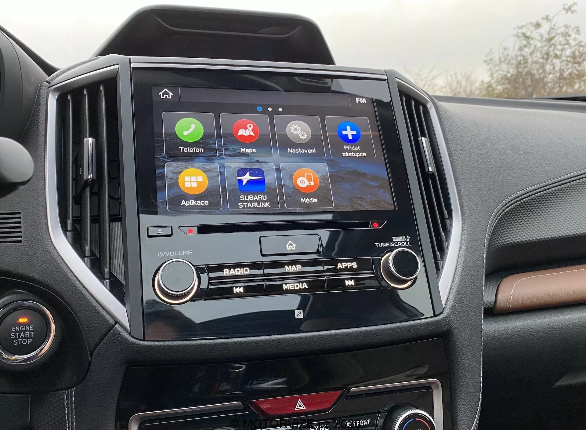 Slika prikazuje sredinsko konzolo Subaru Forester 2020, ki poudarja infotainment sistem in notranjost. V središču slike je zaslon na dotik z ikonami 'Telefon', 'Zemljevidi', 'Trenutno predvaja', 'Sporočila', 'Subaru Starlink', 'Mediji' in 'Aplikacije', kar kaže na različne funkcionalnosti, kot so povezljivost, navigacija in zabava. Infotainment zaslon je v okvirju iz sijajnega črnega materiala, ki kontrastira z dvobarvno armaturno ploščo. Vidni so tudi fizični gumbi in klimatski gumbi, kar ponazarja kombinacijo digitalnih in ročnih kontrol.