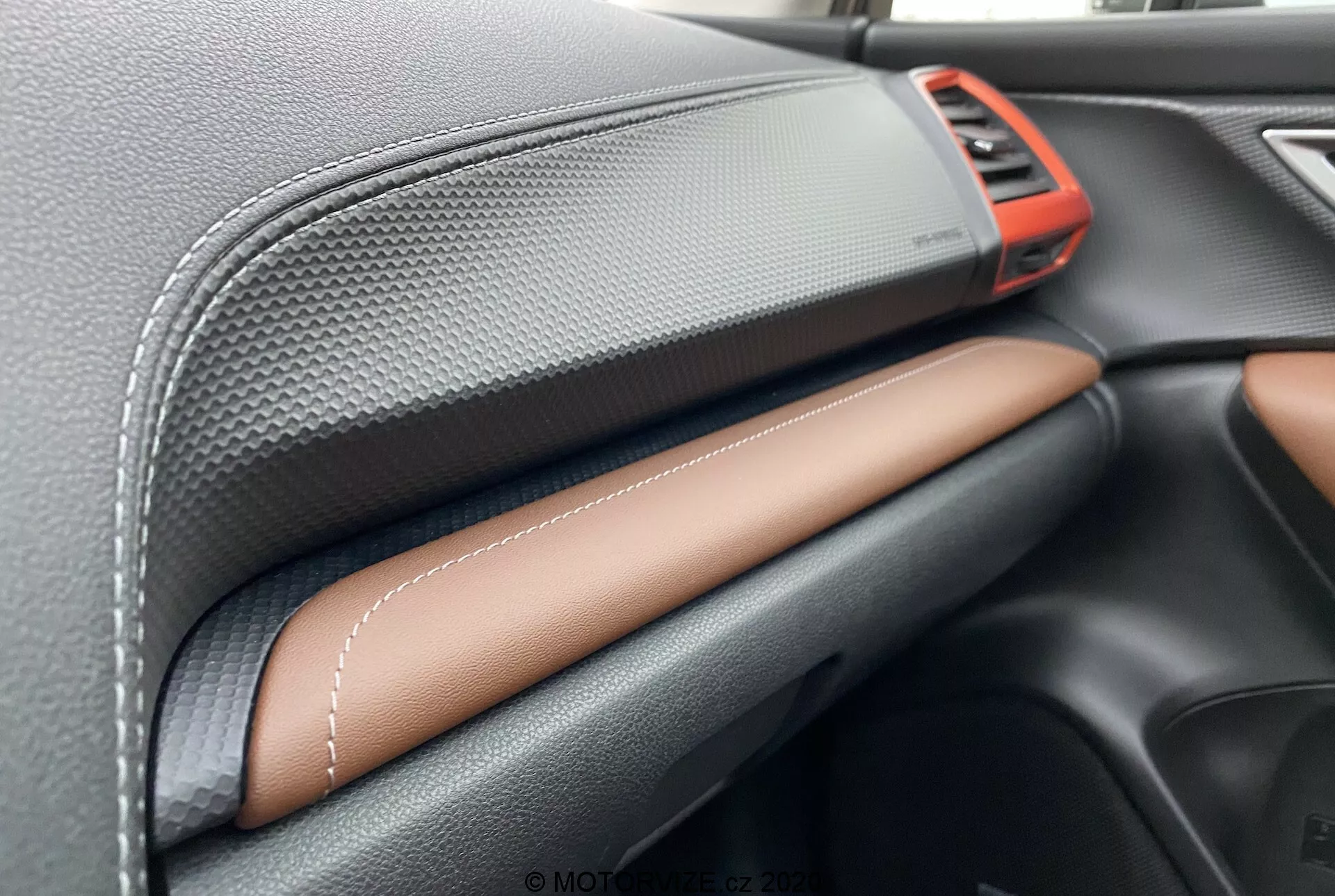 Pogled od blizu na notranjost Subaru Forester 2020, osredotočen na armaturno ploščo. Značilnosti vključujejo svetlo rdeč kovinski zračni ventil v zgornjem desnem kotu, črn material z teksturo ogljikovih vlaken na zgornjem delu armaturne plošče, umetno usnje s kontrastnimi šivi v svetlo rjavi ali rjavi barvi na sredini in eleganten črn predal za rokavice spodaj. To prikazuje obrtniško izdelavo in kakovost vrhunskih materialov.