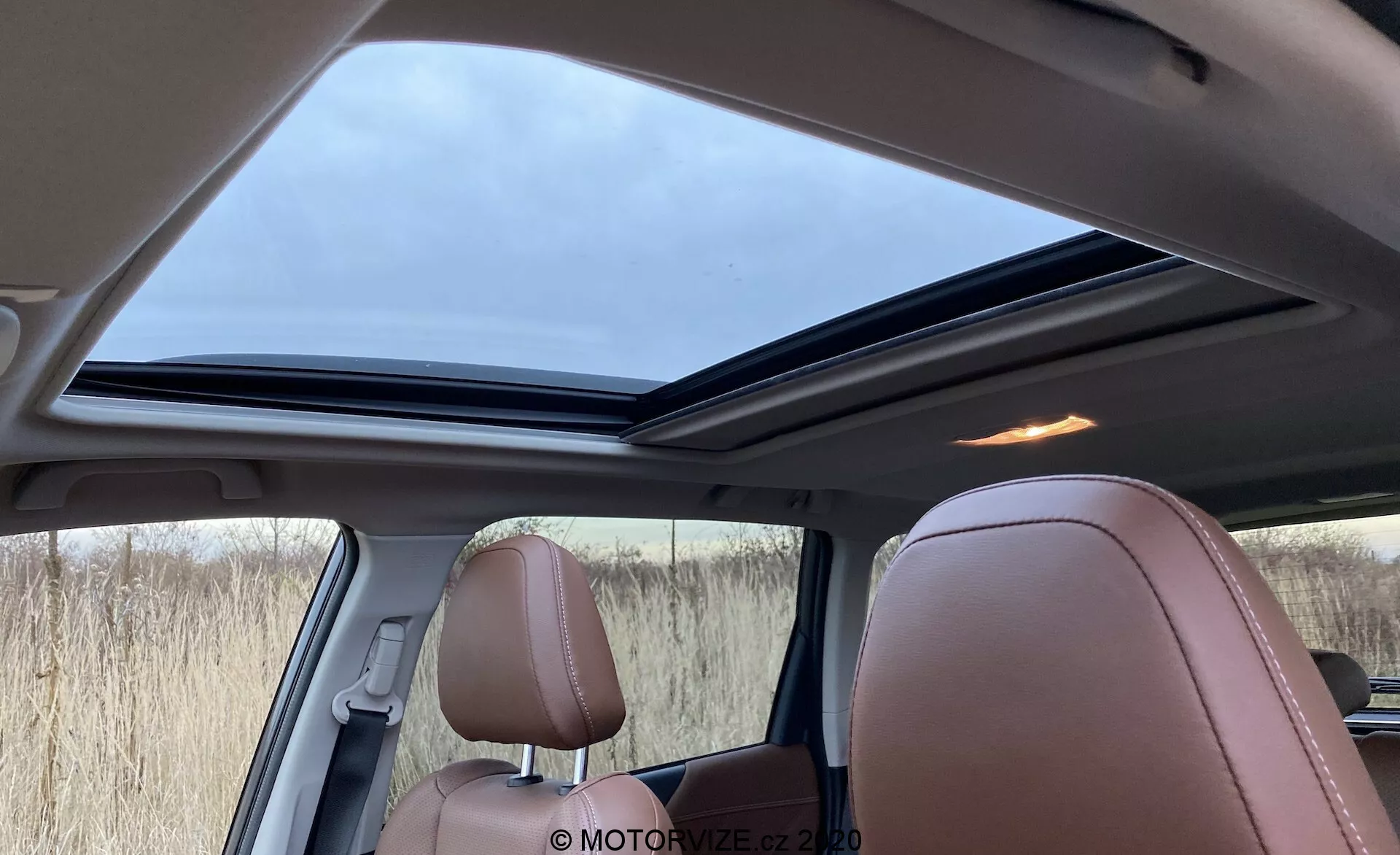 Notranji pogled na Subaru Forester 2020 iz perspektive zadnjega potnika, prikaz velikega panoramskega strešnega okna v rahlo odprtem položaju, razkošnih svetlo rjavih usnjenih sedežev z detajlnim šivom in kombinacije notranjosti v rjavi, črni in bež barvi. Vidna je tudi mrežica zvočnika blizu stebrička in zatemnjena stranska okna, osvetljena z mehko dnevno svetlobo skozi oblačno nebo.