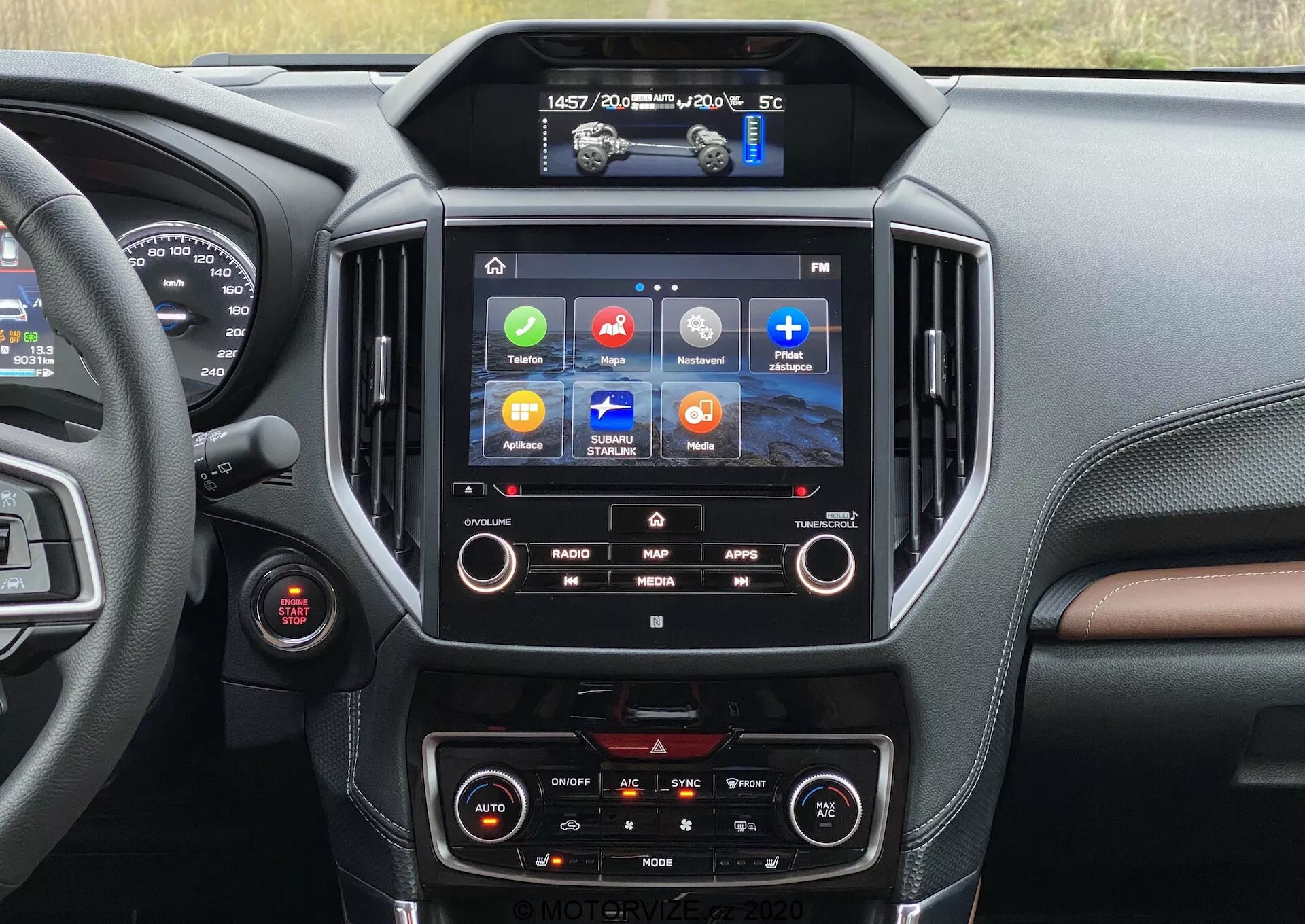 Notranji pogled na Subaru Forester 2020, osredotočen na sredinsko konzolo in infotainment sistem. Zaslon na dotik prikazuje barvne ikone za Radio, Zemljevid, Aplikacije in Medije, kar nakazuje funkcije, kot so GPS navigacija, integracija aplikacij in predvajanje večpredstavnosti. Pod njim so vidni dvopodročne klimatske kontrole. Pogled na voznikov predel razkriva rdeč gumb, verjetno gumb za zagon/ustavitev motorja. Notranji dizajn vključuje temne in rjavkaste materiale, podobne usnju, s šivi, kar kabini daje prefinjen občutek.