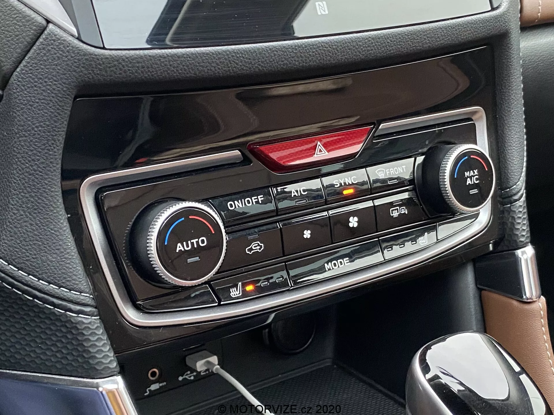 Podroben pogled na sredinsko konzolo Subaru Forester 2020, osredotočena na klimatizacijski panel. Vidni so dvokrožni klimatski upravljalniki z dvema velikima gumboma za nastavitev temperature, gumbi za opozorilne luči, SYNC, MAX AC in sprednji odmrzovalnik vetrobranskega stekla ter LED prikazovalna plošča (neosvetljena). Spodaj so dodatni upravljalniki za smer pretoka zraka, hitrost ventilatorja, kroženje zraka, zadnji odmrzovalnik in vklop/izklop AC, vse na ozadju, ki spominja na ogljikova vlakna. Vidna je tudi del prestavne ročice in držala za pijačo/shranjevalna pregrada.