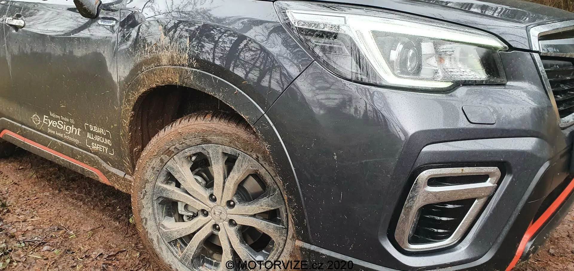 Pogled na sprednji levi del blatnega Subaru Forester 2020, posnet iz nizkega kota, poudarja njegovo oddaljenost od tal, zapleteno večkrako lito platišče, zasnovo spodnjega odbijača in kotne izreze rešetke. Temno obarvano vozilo, verjetno črno ali temno sivo, kaže znake uporabe na terenu z brizganjem blata na sprednjem kolesu, stranskem podboju in stopnici. Pomembne lastnosti vključujejo vlečno kljuko, vgrajeno v odbijač, kovinsko ploščo za zaščito podvozja z rdečo obrobo in omembo 'EyeSight', kar nakazuje na napredne funkcije podpore za voznika.