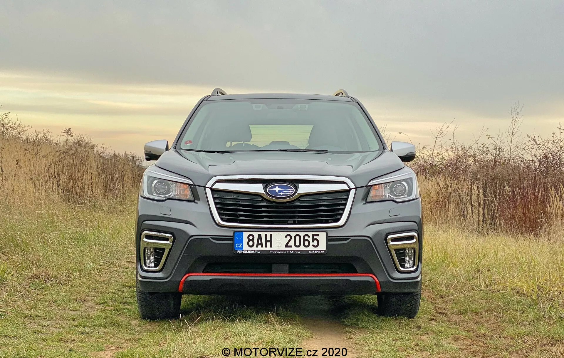 Pogled od spredaj na SUV Subaru Forester 2020, ki prikazuje svojo izpostavljeno rešetko z osrednjo značko logotipa, LED žaromete, meglenke na spodnjem odbijaču in temno sivo sijajno ali kovinsko obdelavo. Simetrična kompozicija poudarja elegantne oblikovalske elemente avtomobila, vključno z morebitnimi parkirnimi senzorji, zračniki in kazalniki ravni opreme. Vozilo je parkirano na zemeljskem površju proti naravnemu ozadju z oblačnim nebom in suho travo.