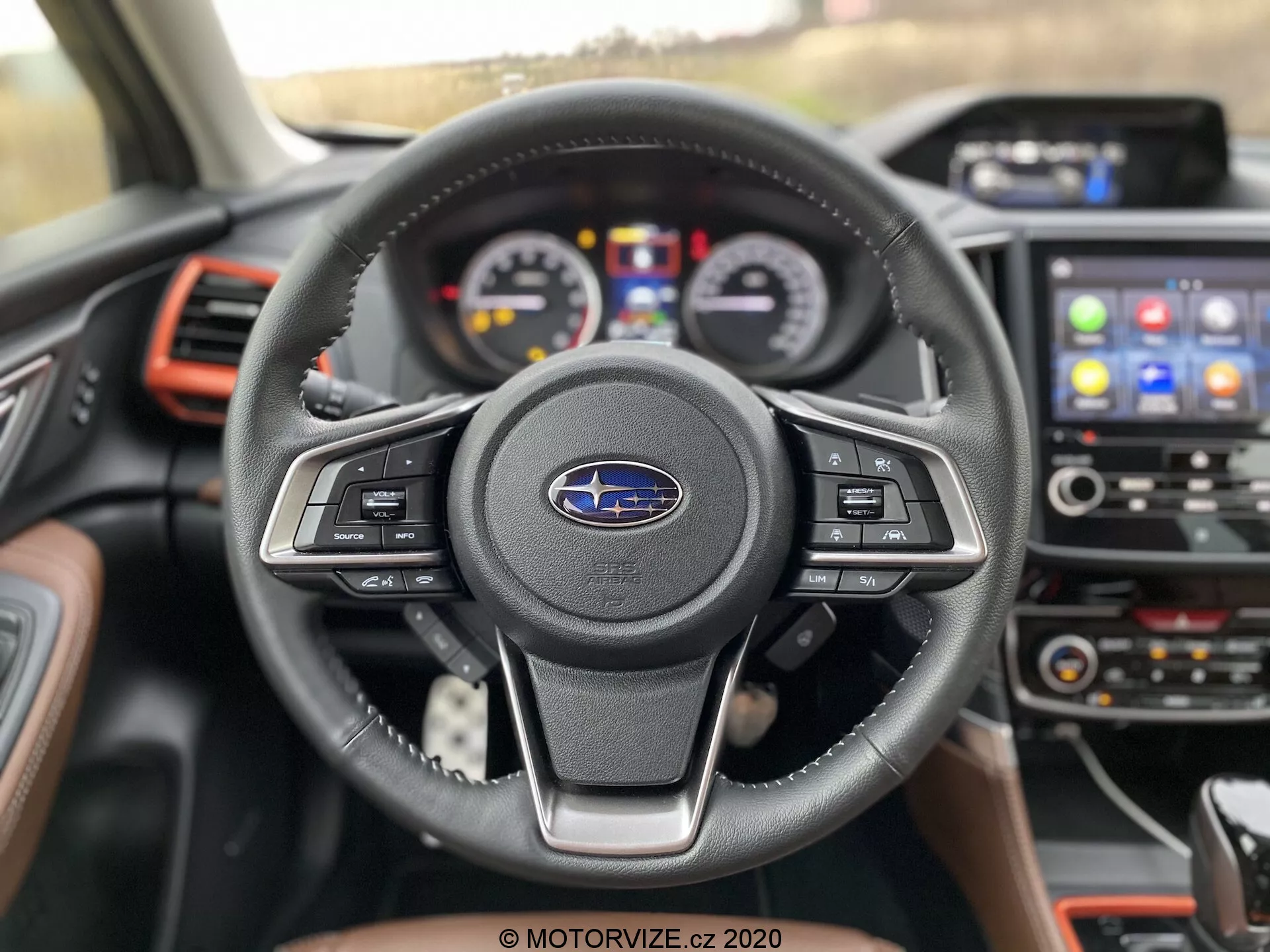 Pogled od blizu na notranjost Subaru Forester 2020 s sedeža voznika, s poudarkom na usnjenem trikraku volanu z večfunkcijskimi kontrolnimi gumbi in emblemom Subaru. Za volanom je viden instrumentna plošča z analognimi številčnicami in osrednjim digitalnim zaslonom. Sredinska konzola vsebuje informacijsko-razvedrilni sistem z zaslonom na dotik, ki prikazuje ikone za navigacijo in povezljivost telefona. Rdeči poudarki na prezračevalnih odprtinah in armaturni plošči dodajo športni pridih sodobnemu in dobro opremljenemu vozilu.