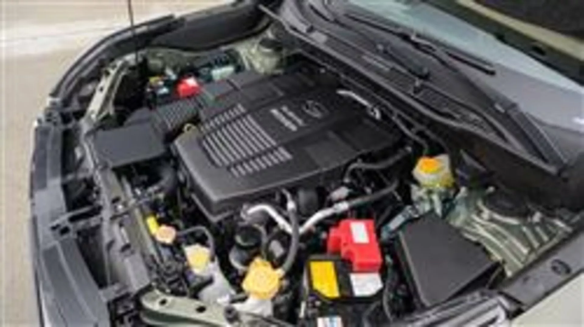 Podroben pogled na motorni prostor Subaru Foresterja 2015 z voznikove strani, ki prikazuje pokrov motorja z znamko, posodo za hladilno tekočino z rumenim pokrovčkom in rdečo prevleko pozitivnega pola baterije med drugimi mehanskimi in električnimi komponentami