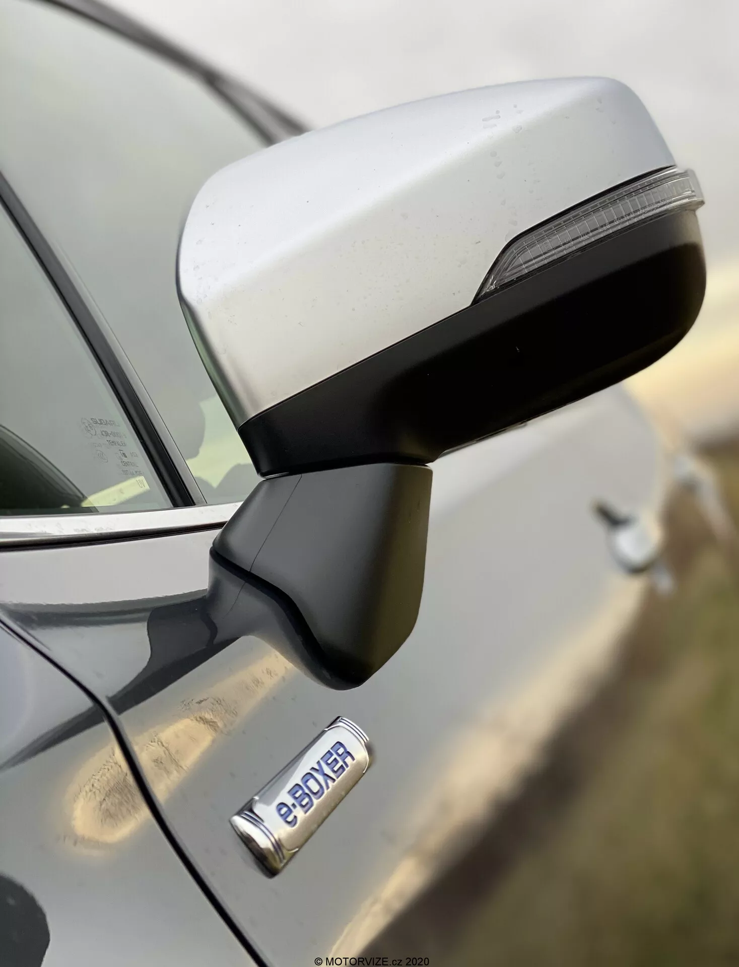 Bližnji posnetek stranskega ogledala in prednjih vrat Subaru Forester 2020 s stranskega pogleda, poudarjen aerodinamičen dizajn ogledala z integriranimi LED smerniki ter kromirano kljuko vrat z gumbom za brezkontaktni vstop. Znak 'e-POWER' nakazuje hibridni ali električni pogonski sklop vozila. Visokoločljivostna slika z majhno globino polja, ki poudarja ogledalo in znak, medtem ko je ozadje zabrisano.