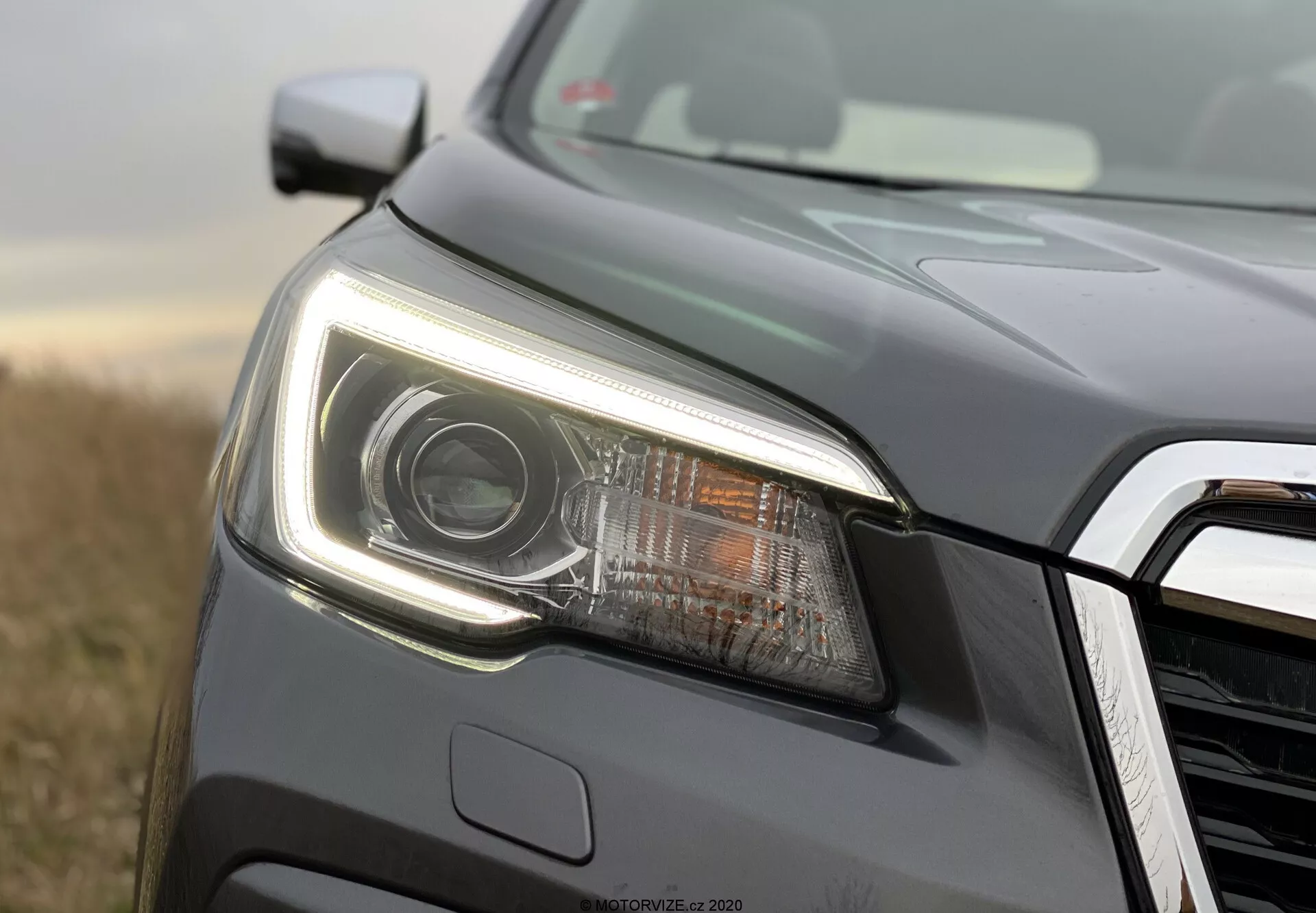 Povečava leve sprednje strani Subaru Foresterja 2020, osredotočena na sodobno ohišje žarometov. Slika, posneta iz nizkega kota, prikazuje izrazit žaromet s projektorsko lečo in LED osvetlitvijo, vključno z ostrimi dnevnimi lučmi. Ohišje žarometa je elegantno s kotnimi linijami in aerodinamično obliko. Zasledi se del sprednje rešetke s kromiranim ali kovinskim zaključkom. Vozilo, pobarvano z metalno temno sivo barvo, je na prostem z naravno svetlobo in zamegljenim rastlinskim ozadjem, ki poudarja podrobnosti žarometa in karoserije s selektivnim fokusom.
