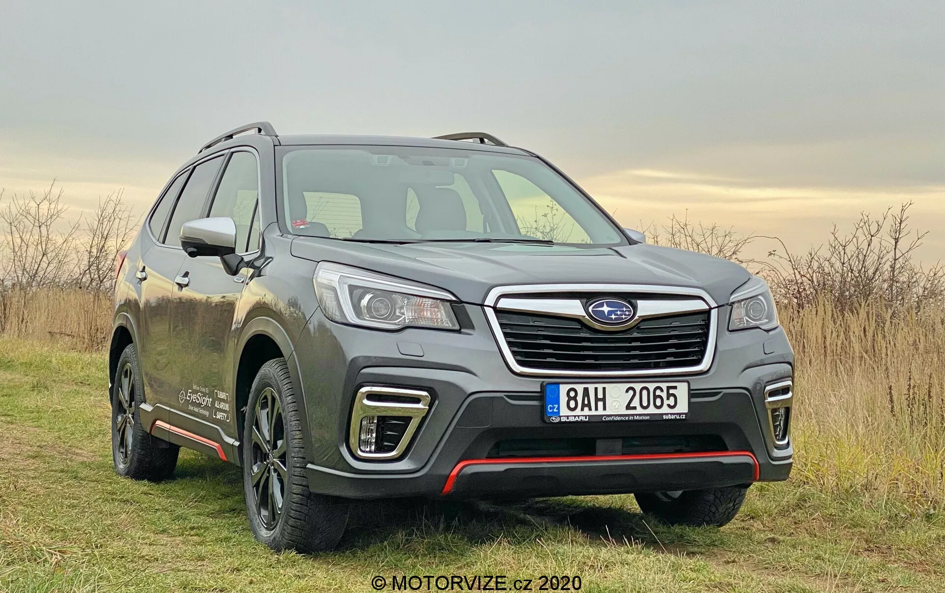 Subaru Forester 2020 na sliki, sodoben SUV, zajet iz prednjega tristranskega kota. Nameščen rahlo desno od okvirja na neravni poti s travo, pod meglennim nebom. Slika poudarja ključne lastnosti, kot so sprednja fasada, mreža z kromirano vodoravno letvico in modulom senzorja, podrobne LED žaromete, konturirane linije pokrova motorja in profil voznikove strani. Vidna lita platišča z večkrakinim dizajnom, ogledala z vgrajenimi smerniki in linije karoserije vzdolž vrat nakazujejo na višjo raven obloge. Blato na vozilu kaže na njegovo uporabo na terenu in makadamskih cestah. Mehka dnevna svetloba in oblačni vremenski pogoji zagotavljajo enakomerno osvetlitev, kar poudarja zasnovo in zmogljivosti vozila.