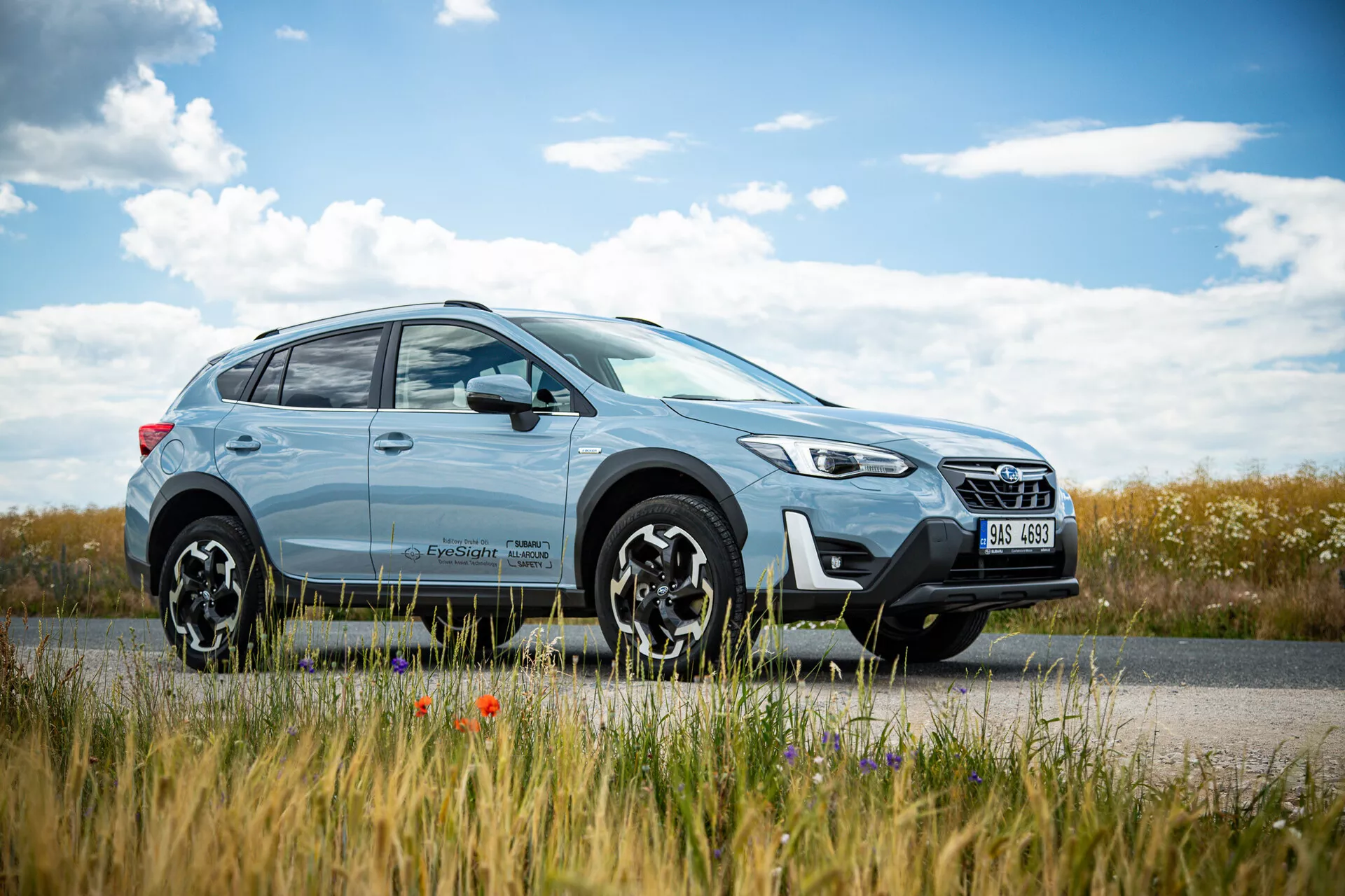 Svetlo modri križanec SUV Subaru XV 2021, parkiran ob cesti z podeželsko pokrajino v ozadju, z modernim dizajnom z LED žarometi in večkrakimi zlitinskimi kolesi, poudarja mestne in terenske sposobnosti.