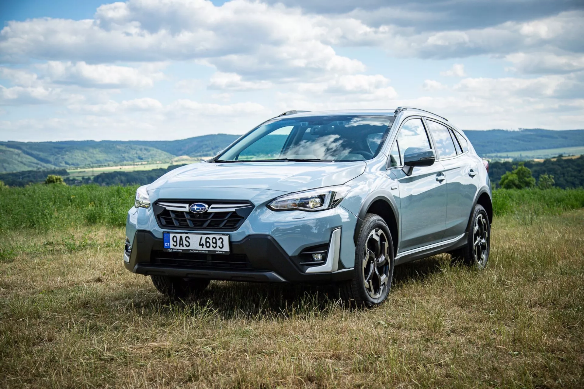 Crossover SUV Subaru XV 2021, parkiran na travi z gričevnatim ozadjem, ki poudarja sprednjo fasado, stranski profil in obliko kolesa