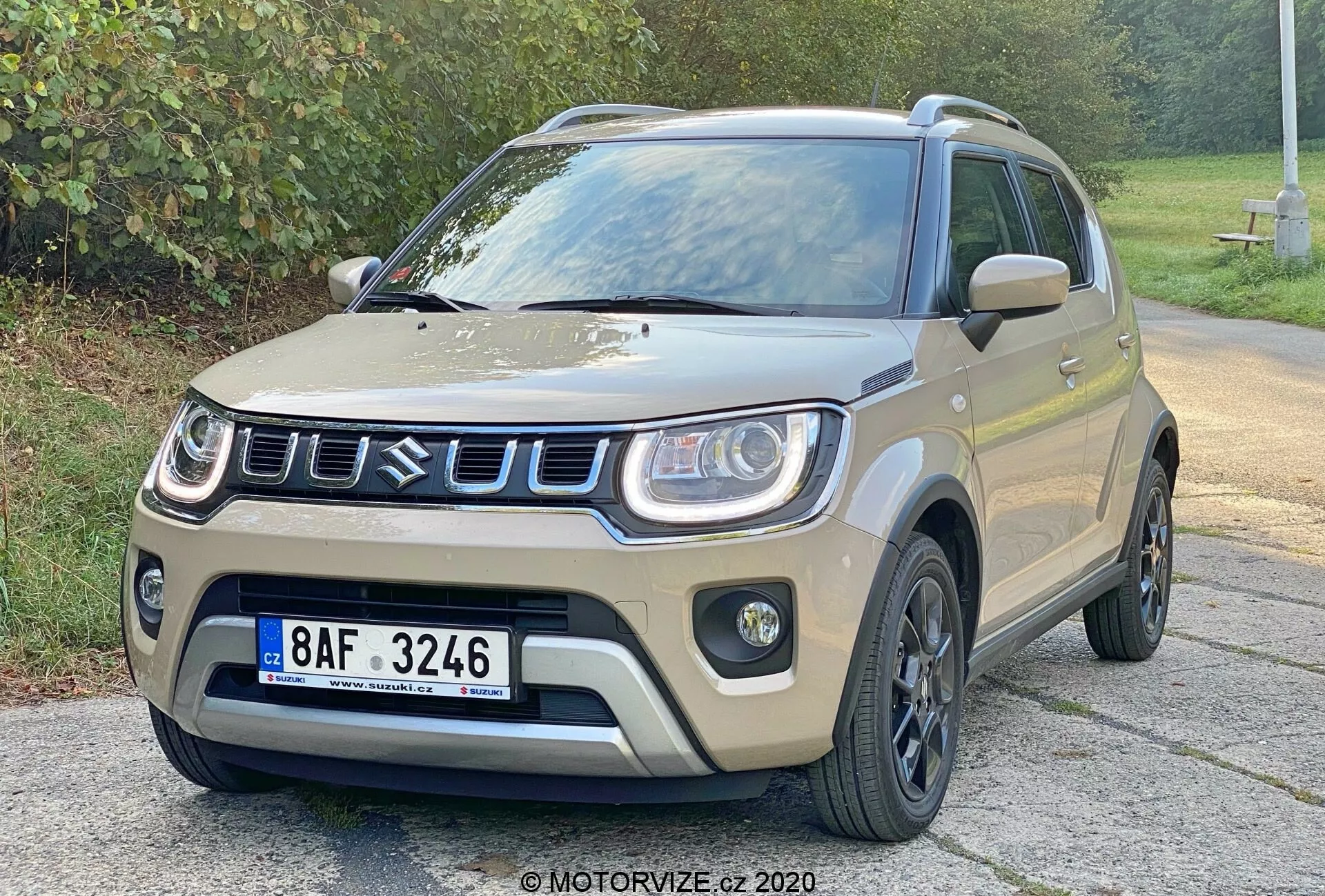 Kompaktni SUV Suzuki Ignis 1.2 Dualjet Hybrid Club iz leta 2020, prikazan iz prednjega tričetrtinskega kota, ki prikazuje njegovo svetlo bež ali peščeno barvo karoserije s črnimi poudarki na blatnikih, spodnjem delu stranskega telesa in strehi. Sprednja maska ima pravokotni vzorec s srebrnim ali kromiranim robom. Sprednji odbijač ima izrazita ohišja meglenk in dinamično obliko pokrova motorja z rahlimi konturami. Črna večkraka lita platišča in črna stranska ogledala dopolnjujejo robusten videz. Vozilo je parkirano na asfaltni površini z zelenim listjem v ozadju pod mehko svetlobo.