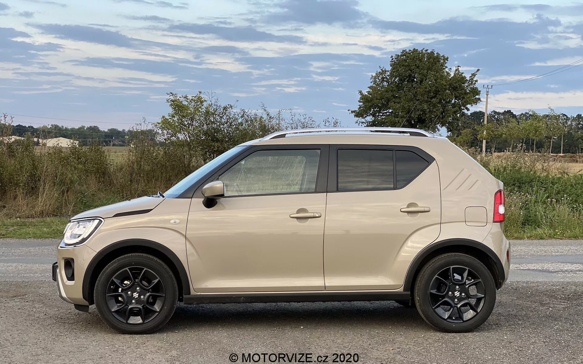 Stranski profil kompaktnega SUV-ja Suzuki Ignis 1.2 Dualjet Hybrid Club 2020 s štirimi vidnimi vrati, črnimi petkrakimi lita platišči, dvobarvnim bež in črnim dizajnom, zatemnjenimi okni in možnostjo integriranih ogledal z utripalkami v podeželskem okolju.
