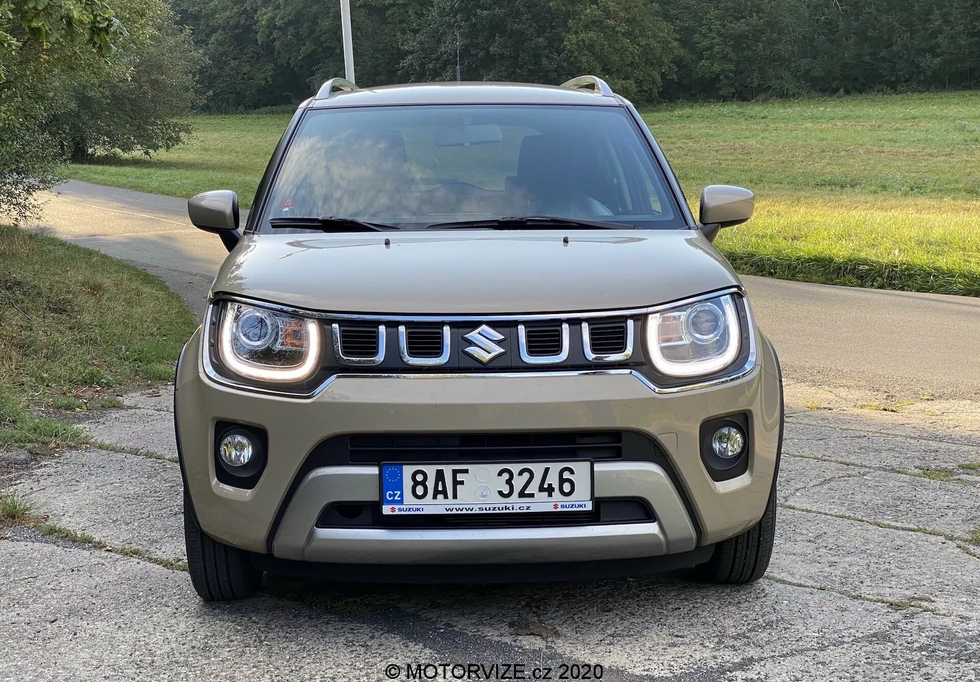 Pogled spredaj na kompaktni SUV Suzuki Ignis 2020 na tlakovani cesti s travnatim ozadjem, podeželsko ali parkovno okolje, posnetek iz nizkega kota poudarja sprednji dizajn, z LED dnevnimi lučmi, izrazito masko z emblemom, robustnim sprednjim odbijačem z meglenkami, mat barva v zemeljskem zeleno-rjavem odtenku